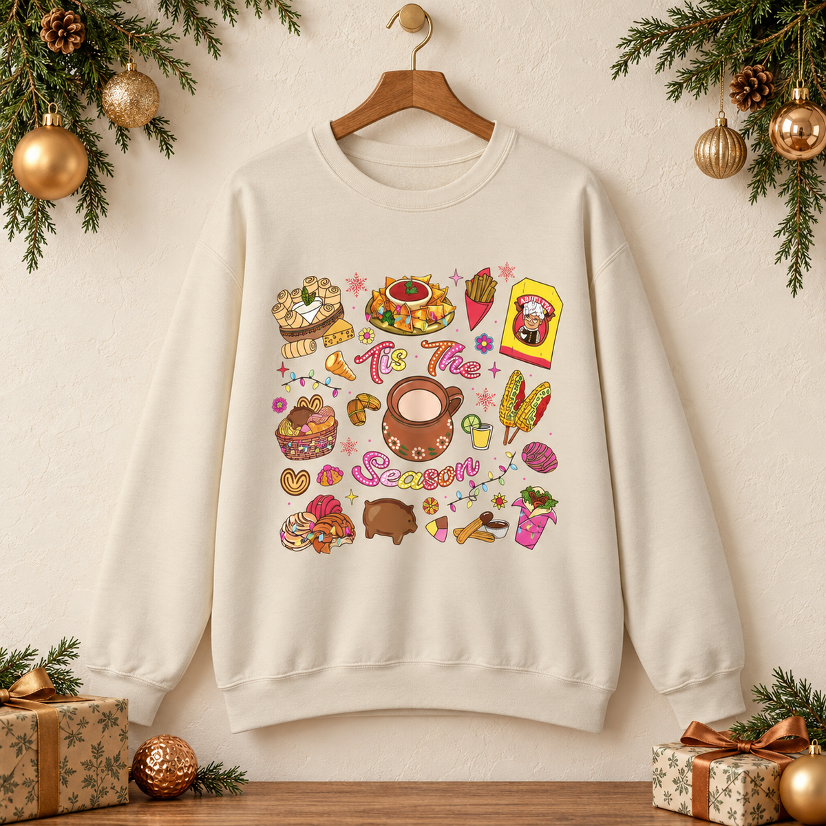 An beige crewneck sweatshirt hanging on a wooden hanger against a warm beige wall, decorated with Christmas greenery, ornaments, and wrapped gifts. The crewneck sweatshirt features a colorful Christmas-themed graphic print with illustrations of seasonal hispanic treats and the text 'Tis The Season'.