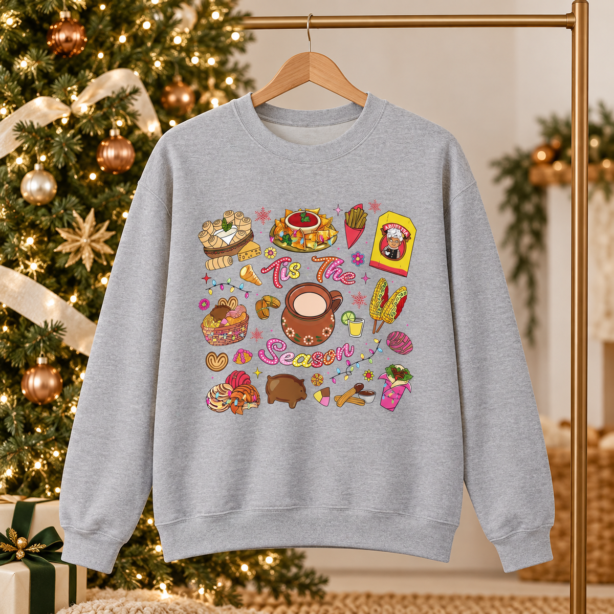 A light gray crewneck sweatshirt featuring a colorful Christmas-themed graphic print with illustrations of seasonal hispanic treats and the text 'Tis The Season', in front of a decorated Christmas tree.