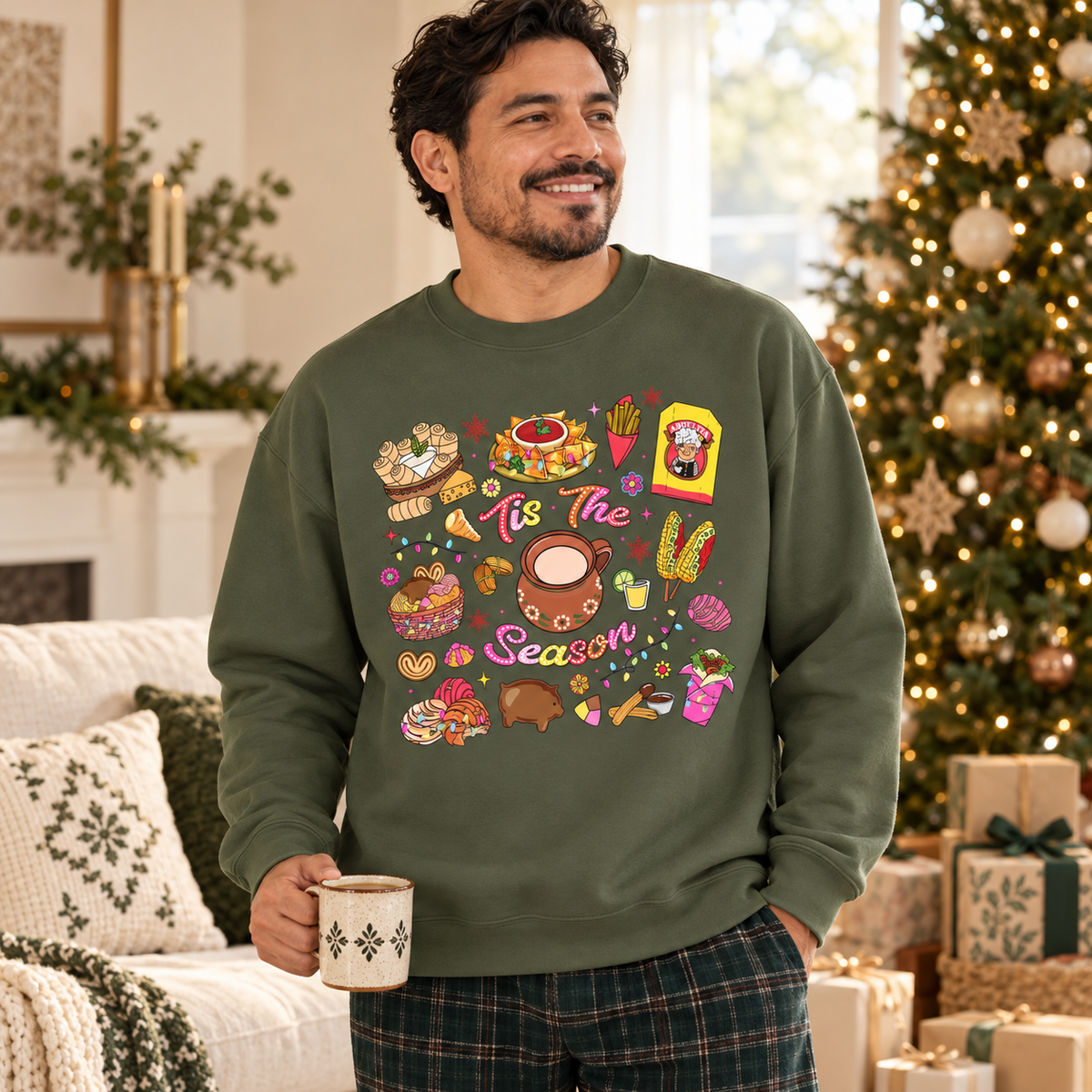 A man wearing an olive green crewneck sweatshirt featuring a colorful Christmas-themed graphic print with illustrations of seasonal hispanic treats and the text 'Tis The Season', surrounded by festive illustrations. He is holding a mug in a cozy, decorated living room with a Christmas tree and wrapped gifts in the background.