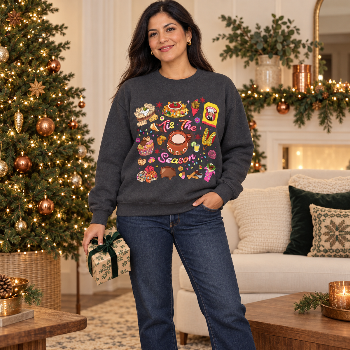 A woman stands in a decorated living room wearing a dark gray crewneck sweatshirt featuring a colorful Christmas-themed graphic print with illustrations of seasonal hispanic treats and the text 'Tis The Season'. She is wearing blue jeans and holding a small wrapped gift box.