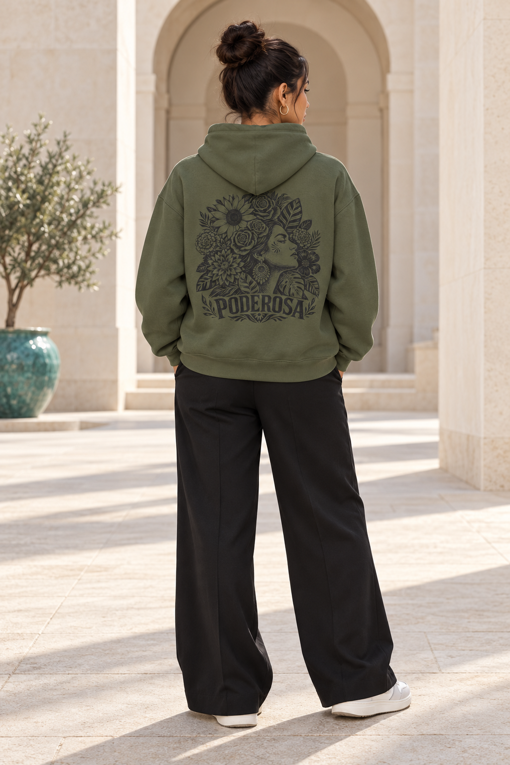 A woman viewed from behind wearing an olive green hoodie with a large black graphic print on the back and black wide-leg pants, standing in a sunlit outdoor walkway with stone columns.