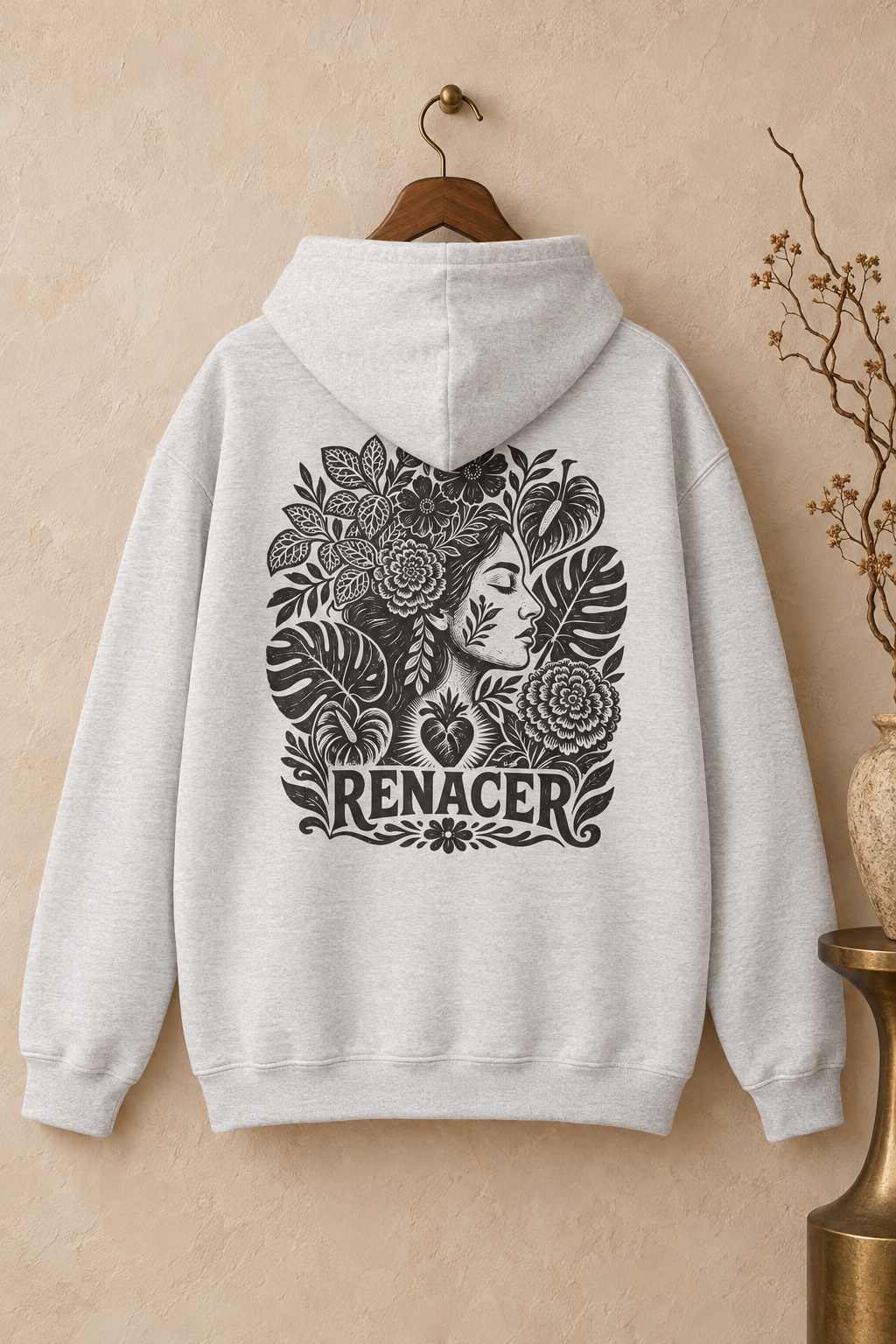 A light gray heather pullover hoodie hanging on a wooden hanger against a textured beige wall. The back of the hoodie features a large black graphic print of a woman's profile surrounded by flowers and leaves, with the word 'RENACER' printed below.