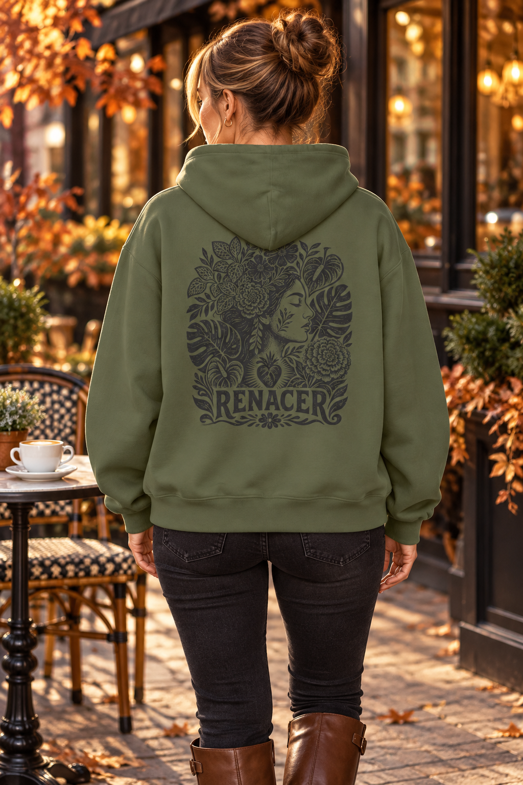 A woman viewed from behind wearing an olive green hoodie with a large dark graphic print on the back featuring a woman's face surrounded by flowers and the word 'RENACER'. She is standing outdoors on a cobblestone street in front of a storefront with warm lights and autumn foliage.