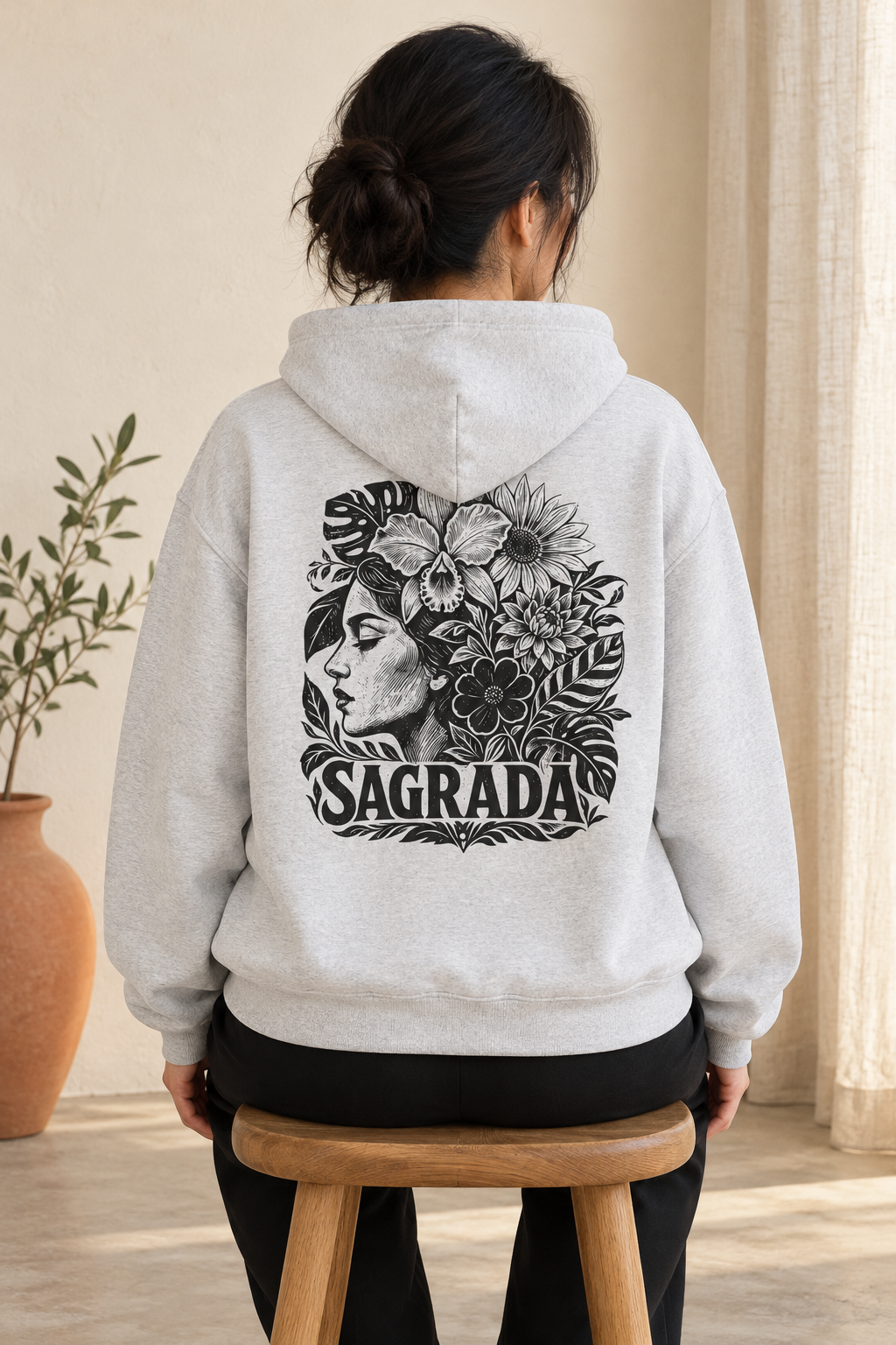 A person viewed from behind wearing a light heather gray hoodie with a large black graphic on the back featuring floral, skull, and profile artwork with the word 'SAGRADA.' The person is seated on a wooden stool in a softly lit indoor setting with curtains and a plant visible.