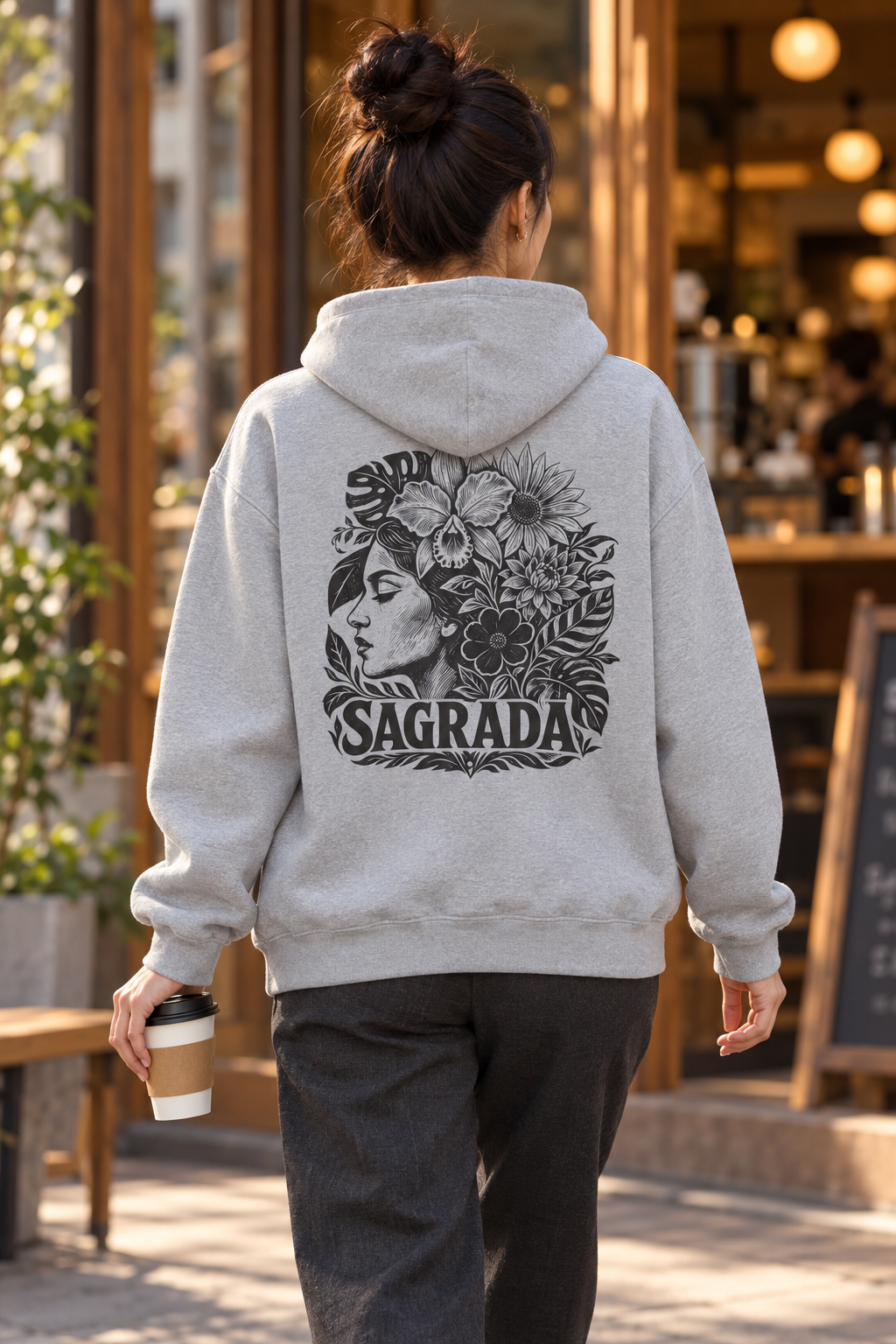 A person viewed from behind wearing a light gray hoodie with a large black graphic print on the back featuring a woman's face, flowers, and the word 'SAGRADA'. The person is walking outdoors in front of a cafe, holding a coffee cup.