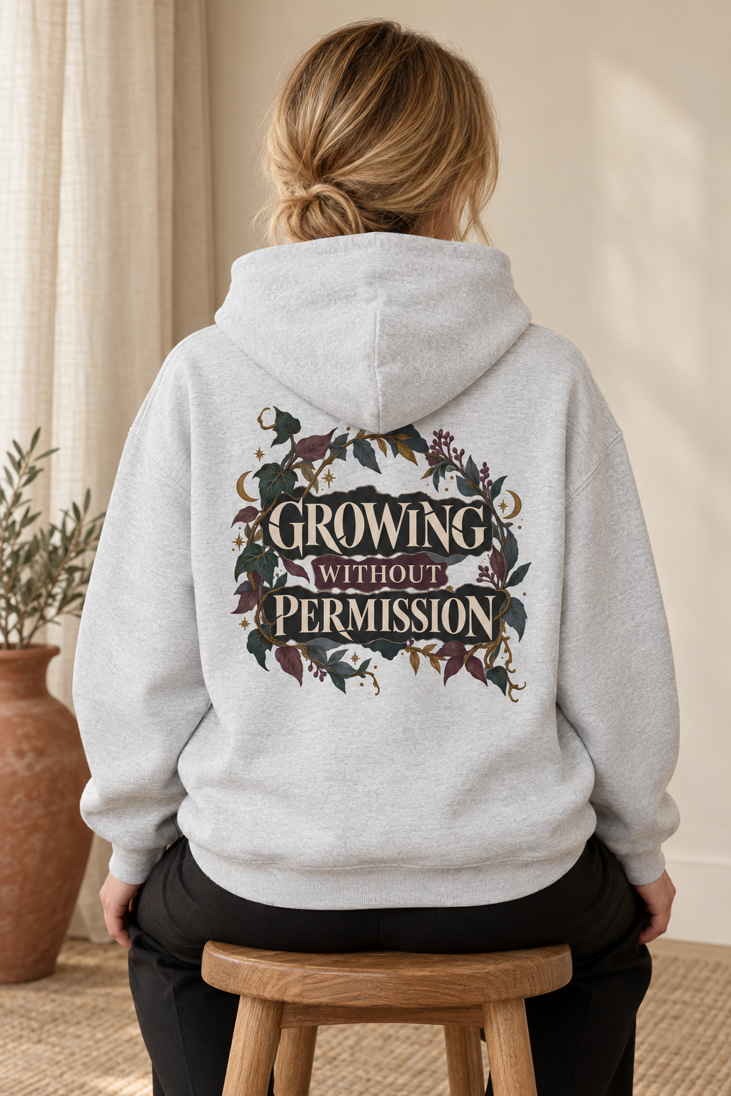 A woman viewed from behind wearing a light heather gray hoodie with a large graphic on the back that reads 'GROWING WITHOUT PERMISSION' in cream lettering surrounded by floral illustrations. She is seated on a wooden stool in a warm indoor setting.