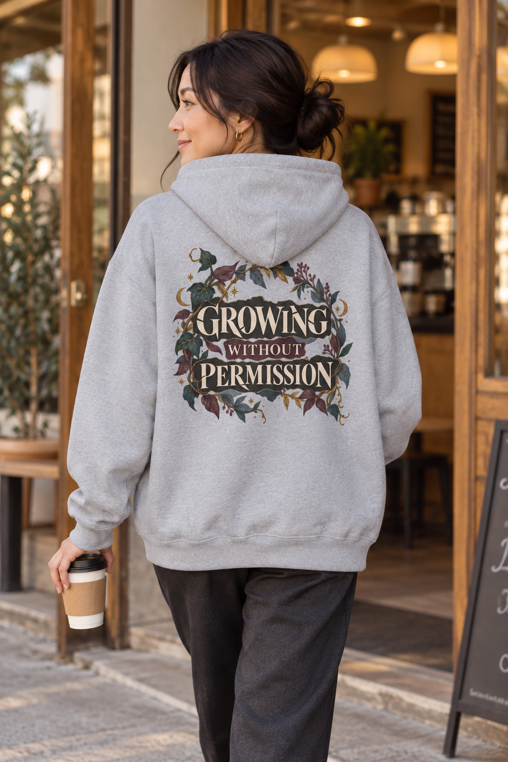 A woman wearing a light gray hoodie with a large graphic on the back that reads 'GROWING WITHOUT PERMISSION' in a floral wreath design, standing in front of a coffee shop.
