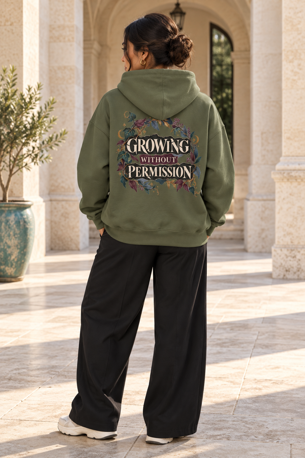 A woman viewed from behind wearing an olive green hoodie with a colorful graphic on the back that reads 'GROWING WITHOUT PERMISSION' in cream lettering, paired with black wide-leg sweatpants in an outdoor stone walkway setting.
