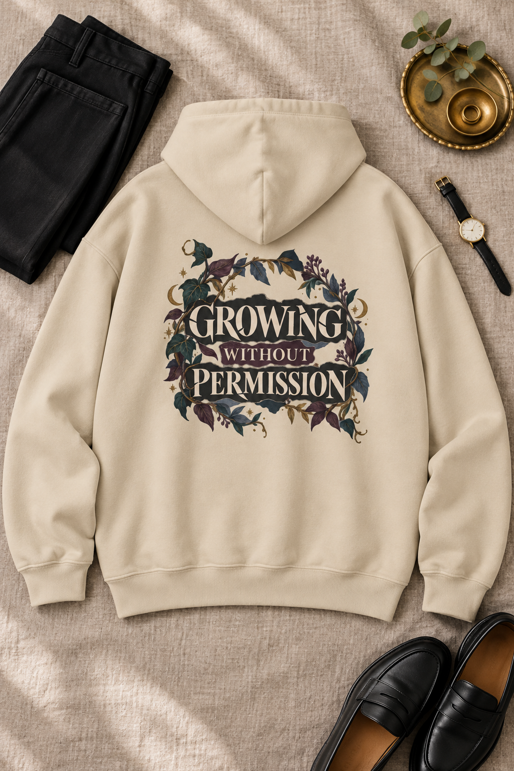 A beige-colored hoodie is laid flat on a textured beige fabric background, styled with black pants, a watch, a small gold tray with greenery, and black loafers. The back of the hoodie features a graphic print with the text 'GROWING WITHOUT PERMISSION' surrounded by dark purple, green and blue floral elements.