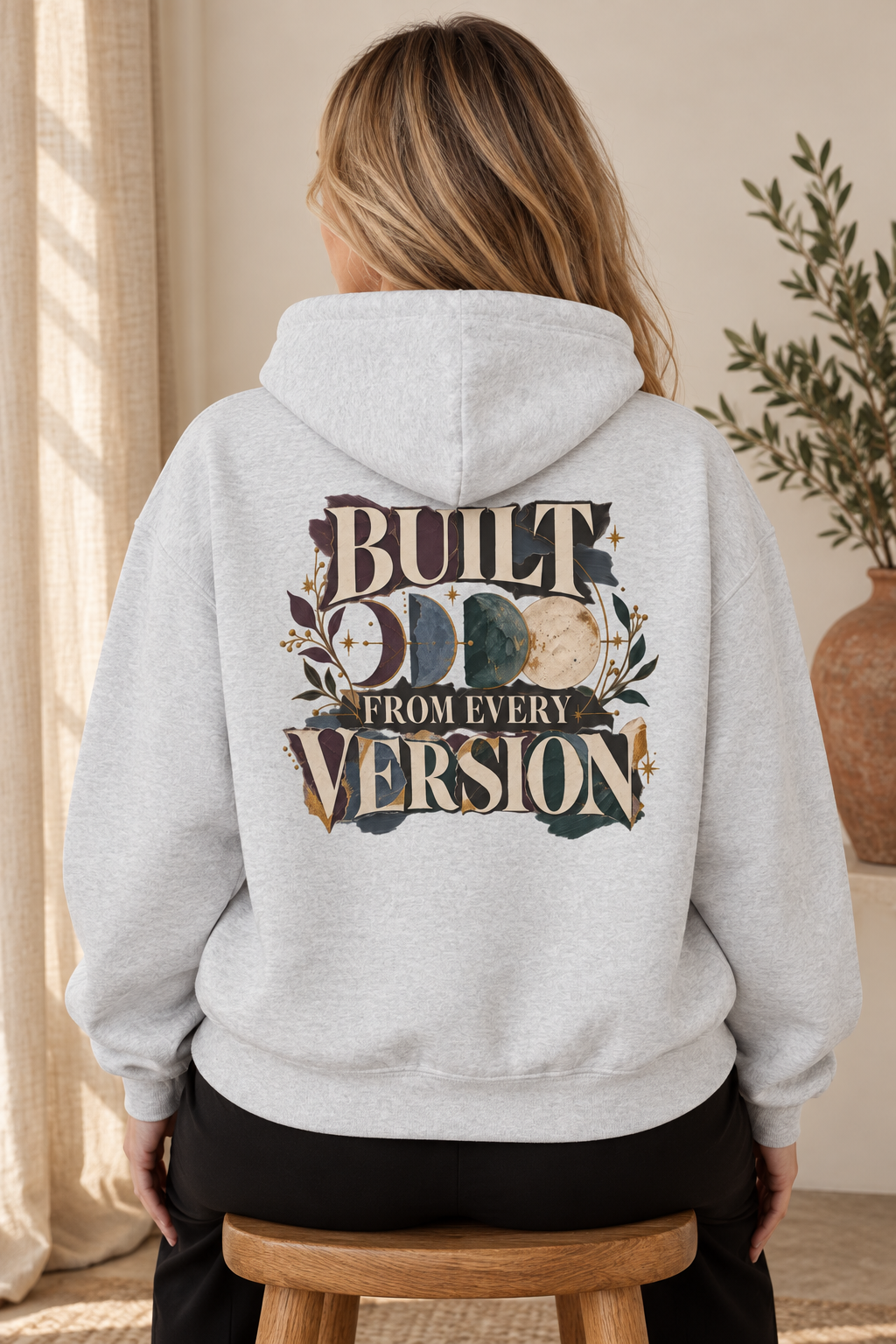 A woman viewed from behind wearing a light heather gray pullover hoodie shown from the back, featuring a large graphic print with the words 'BUILT FROM EVERY VERSION' and a moon phase design in muted earthy tones. She is seated on a wooden stool in a softly lit indoor setting with curtains and a plant visible.