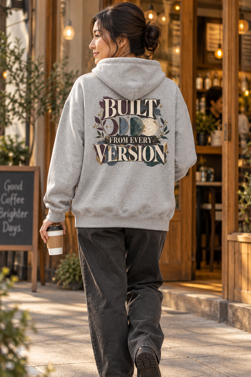 A woman viewed from behind walking on a sidewalk in front of a coffee shop, wearing a light gray hoodie with a large graphic print on the back that reads 'BUILT FROM EVERY VERSION' and dark gray sweatpants.