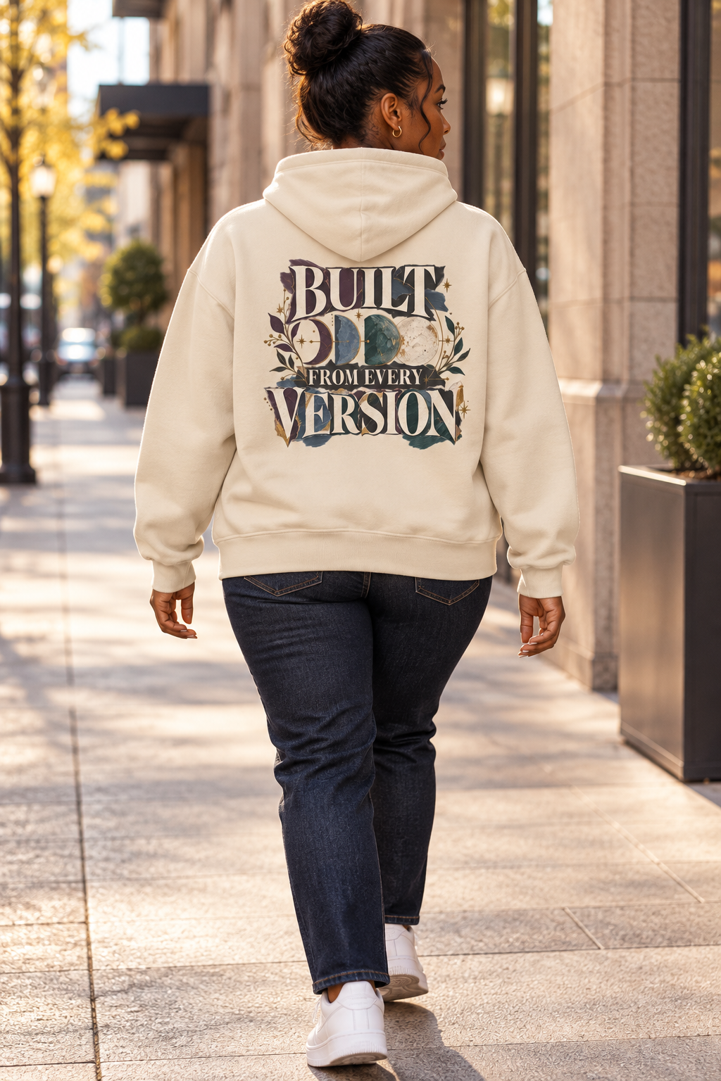 A woman viewed from behind walking on a city sidewalk, wearing a beige-colored hoodie with a large graphic on the back that reads 'BUILT FROM EVERY VERSION' in cream lettering with teal and beige floral details.