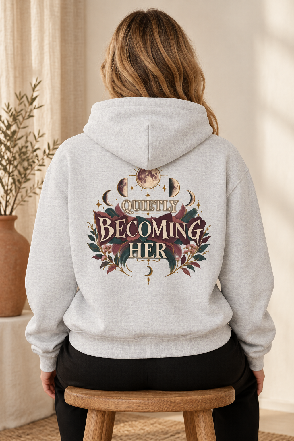 A woman viewed from behind wearing a light heather gray hoodie with a large decorative graphic on the back that reads 'Quietly Becoming Her' with moon phases and floral elements. She is seated on a wooden stool in a softly lit indoor setting.
