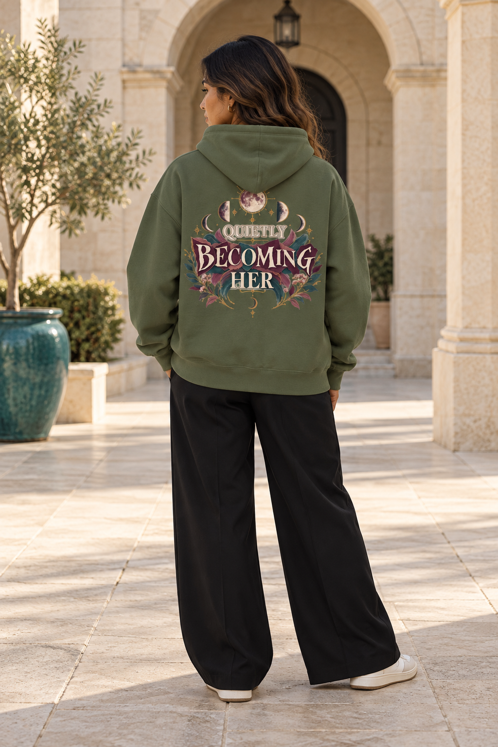 A woman stands outdoors in a stone walkway, viewed from the back, wearing an olive green hoodie with a large graphic on the back that reads 'Quietly Becoming Her' with floral details, paired with black wide-leg pants and white sneakers.