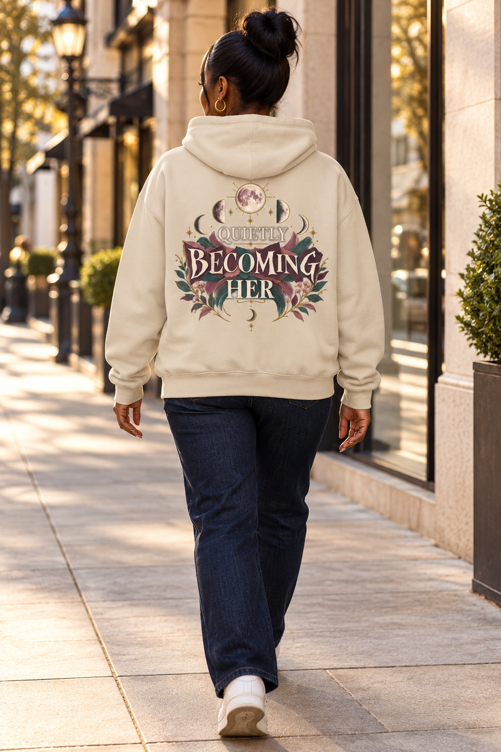 A woman viewed from behind walking on a city sidewalk, wearing a beige-colored hoodie with a large graphic on the back that reads 'BECOMING HER' surrounded by floral and celestial designs, paired with dark blue jeans and white sneakers.
