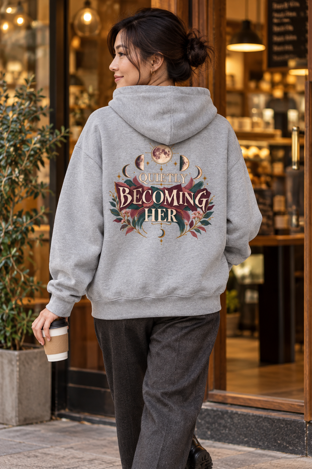 A woman wearing a light gray pullover hoodie shown from the back, featuring a large graphic print with the words 'Quietly Becoming Her' surrounded by decorative floral and celestial motifs in muted maroons, green, and gold tones, standing in front of cafe.