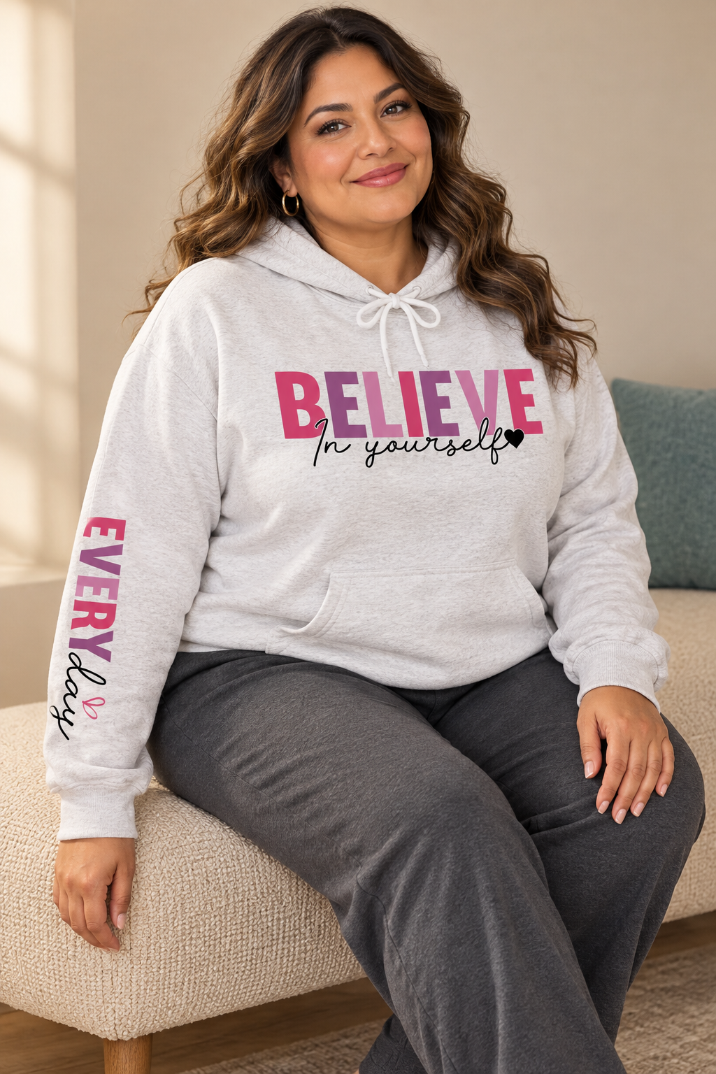 A woman sitting on a beige couch wearing a light heather gray pullover hoodie featuring a graphic on the chest that reads 'BELIEVE In yourself' in different shades of pink and black, with 'EVERYDAY' printed vertically in different shades of pink and black on the left sleeve.