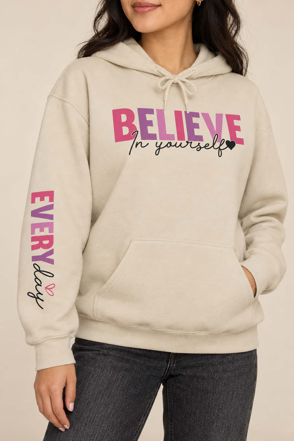 A woman wearing a beige-colored pullover hoodie featuring a graphic on the chest that reads 'BELIEVE In yourself' in different shades of pink and black, with 'EVERYDAY' printed vertically in different shades of pink and black on the left sleeve.