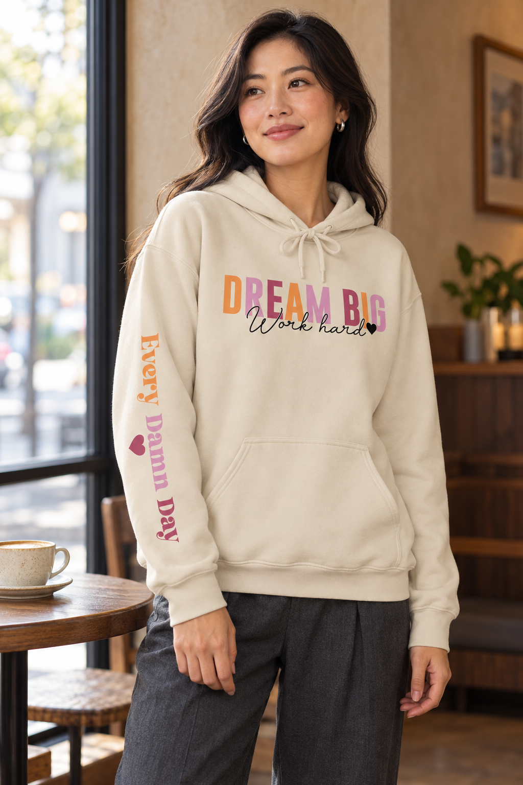 A woman wearing a beige-colored pullover hoodie with a colorful graphic on the chest reading 'DREAM BIG Work hard' and 'EVERY DAMN DAY' with a heart printed vertically on the sleeve,  in a cafe setting.