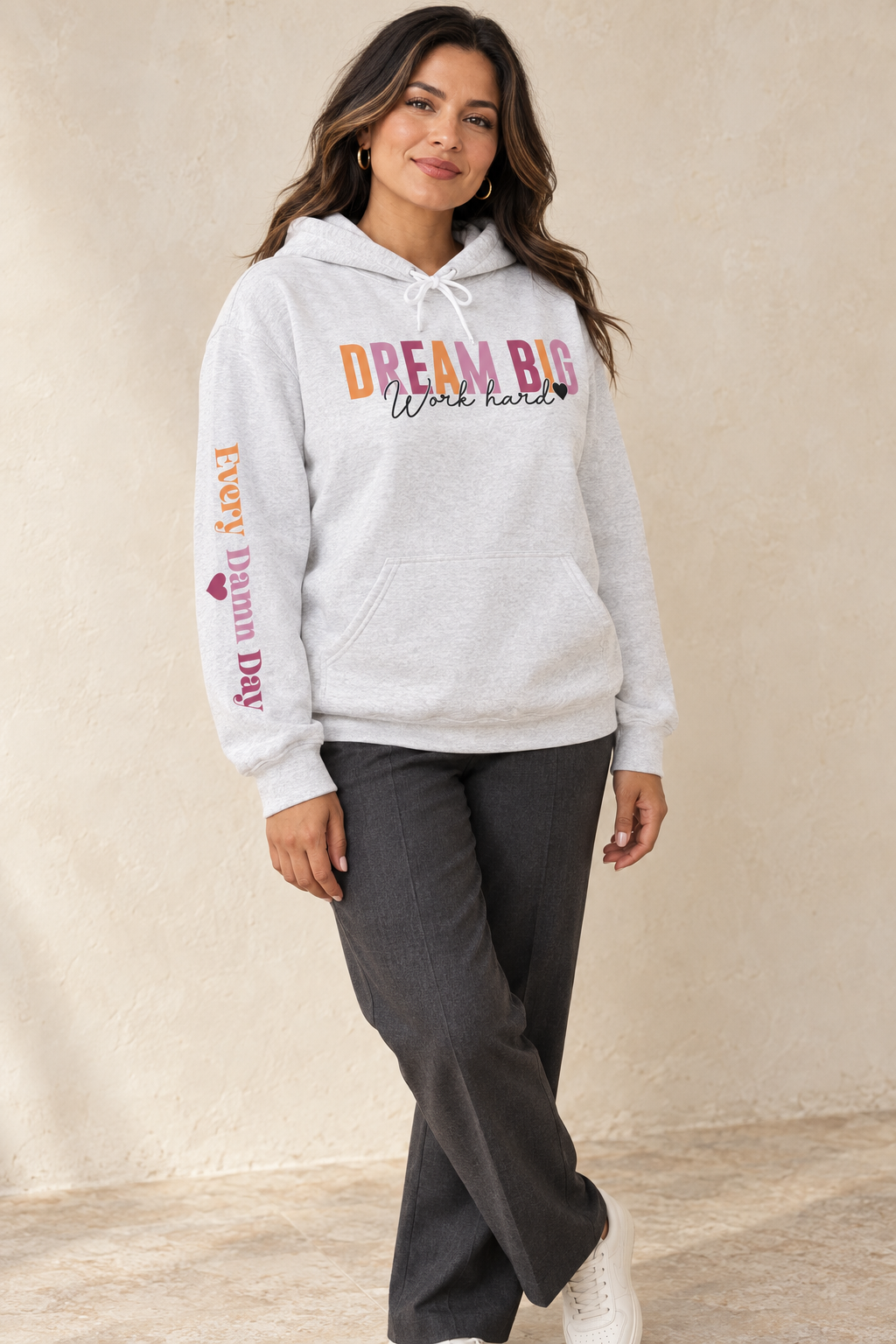 A woman modeling a light gray heather pullover hoodie with a colorful graphic on the chest reading 'DREAM BIG Work hard' and 'EVERY DAMN DAY' with a heart printed vertically on the sleeve, paired with dark gray pants against a textured beige wall.