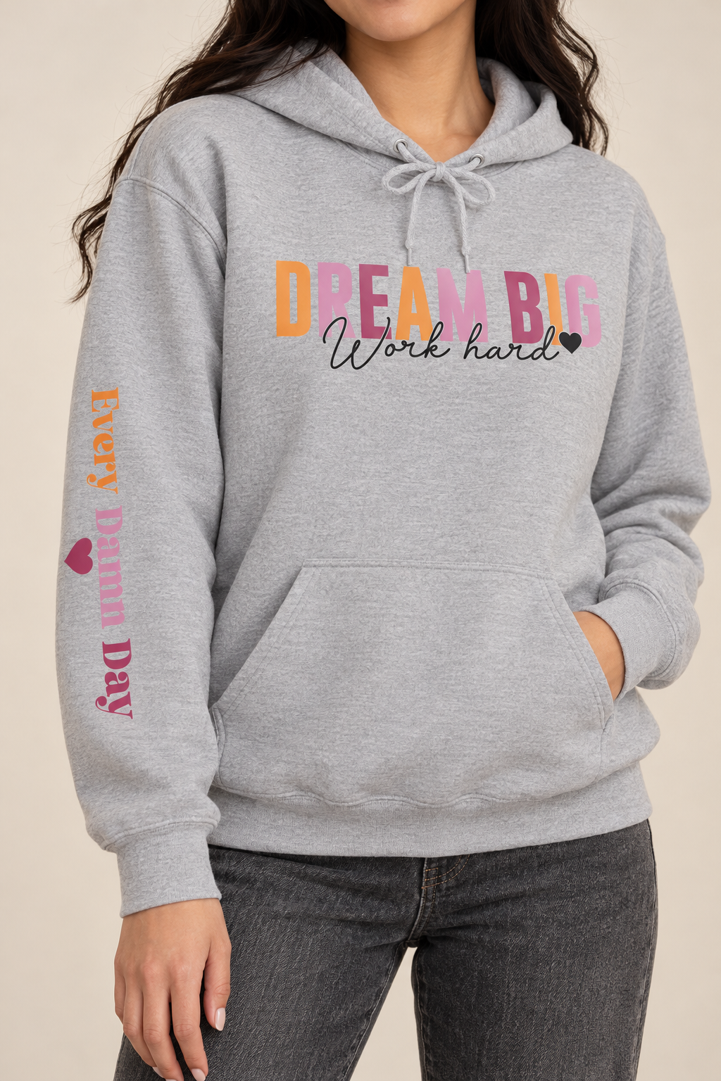 A woman wearing a light gray heather pullover hoodie with a colorful graphic on the chest reading 'DREAM BIG Work hard' and 'EVERY DAMN DAY' with a heart printed vertically on the sleeve.