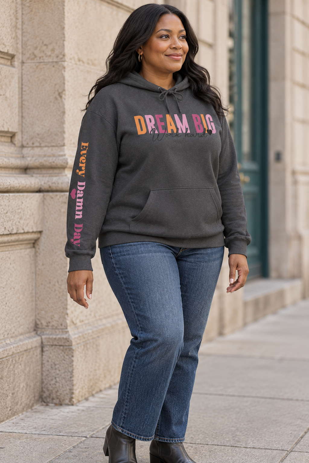 A woman wearing a dark gray pullover hoodie with a colorful graphic on the chest reading 'DREAM BIG Work hard' and 'EVERY DAMN DAY' with a heart printed vertically on the sleeve, paired with blue jeans, standing outdoors against a stone building.