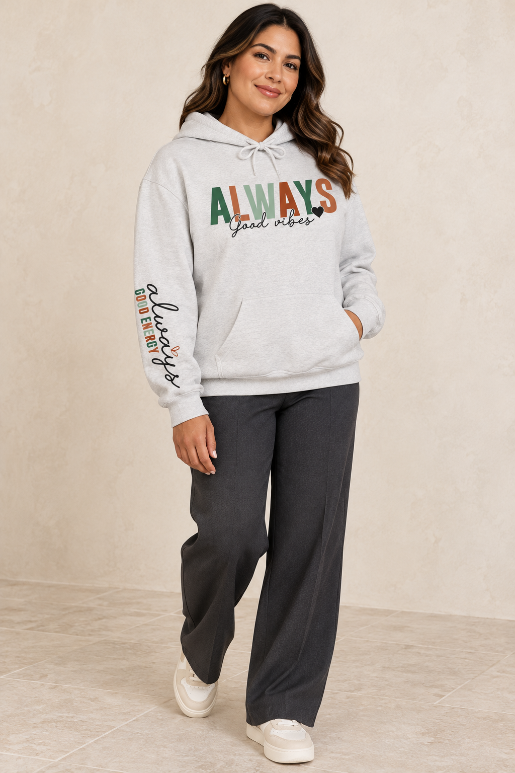 A woman modeling a light gray pullover hoodie with a front kangaroo pocket and drawstring hood, featuring the word 'ALWAYS' in block letters with 'Good vibes' in black script underneath with a solid small heart. The left sleeve has a vertical script graphic 'ALWAYS GOOD ENERGY' paired with dark gray wide-leg sweatpants and white sneakers, set against a neutral beige studio backdrop.
