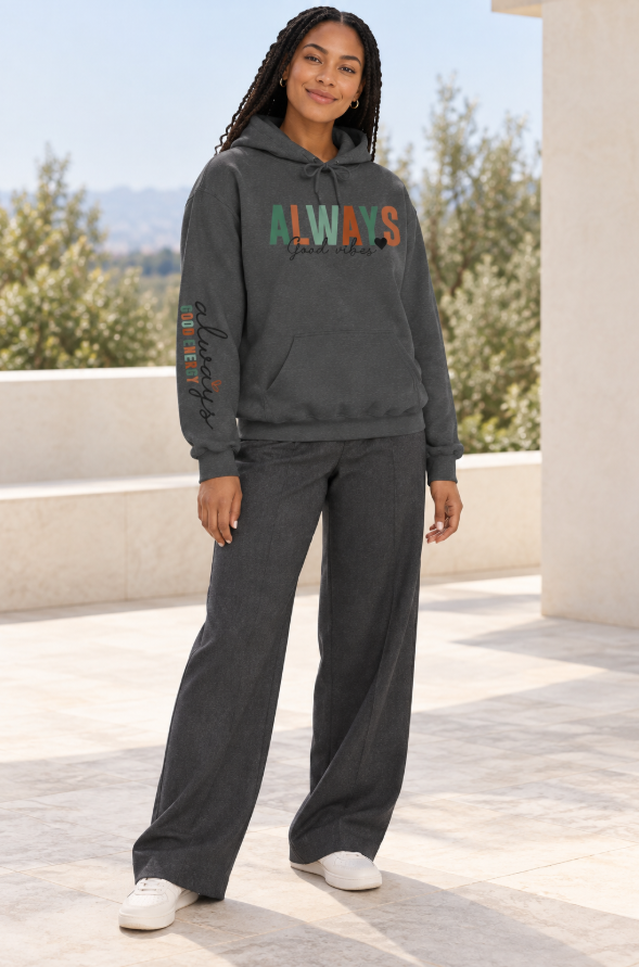 A woman modeling a dark gray pullover hoodie with a front kangaroo pocket and drawstring hood, featuring the word 'ALWAYS' in block letters with 'Good vibes' in black script underneath with a solid small heart. The left sleeve has a vertical script graphic 'ALWAYS GOOD ENERGY' and matching wide-leg sweatpants, standing outdoors on a patio.