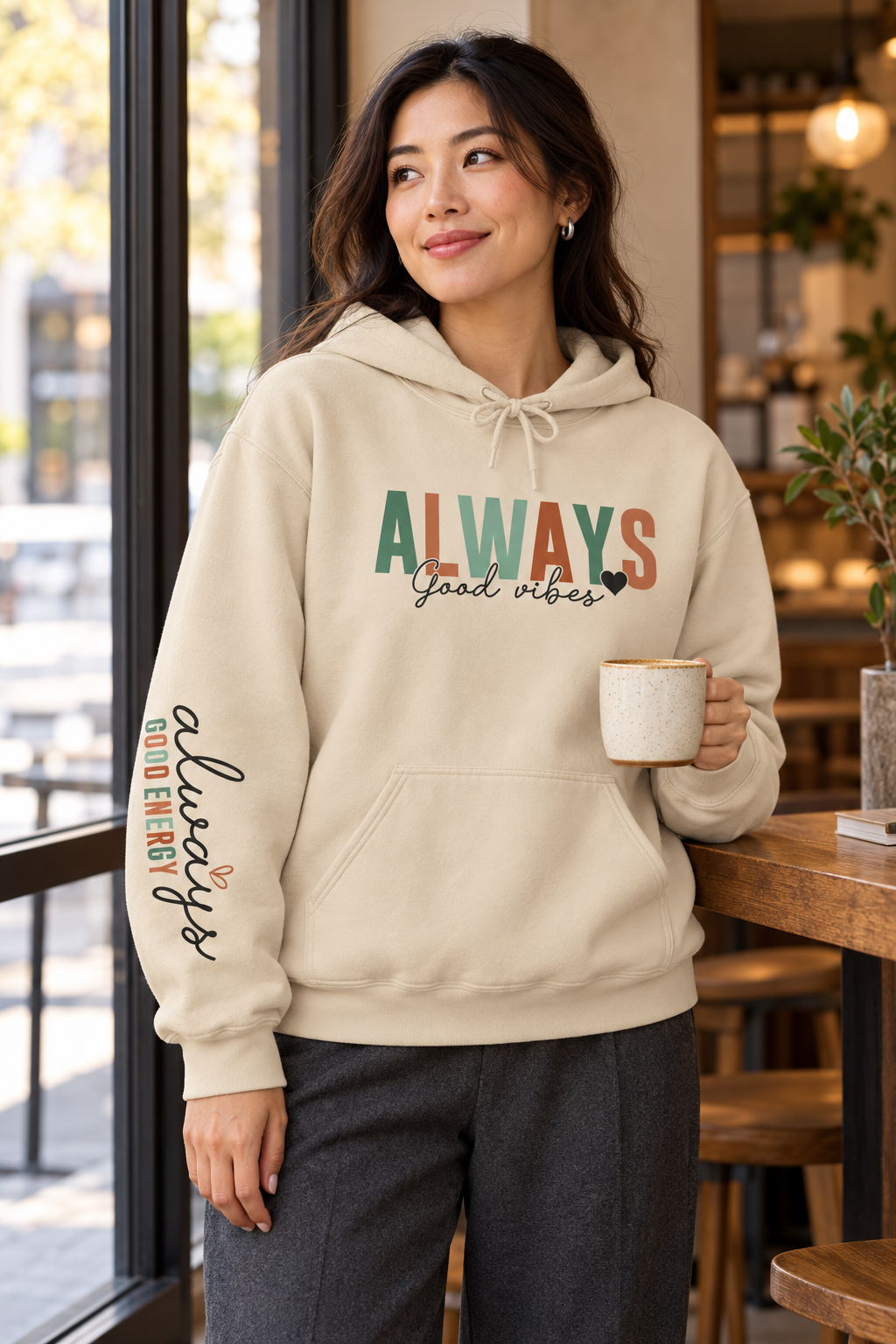 A woman wearing a beige pullover hoodie with a front kangaroo pocket and drawstring hood, featuring the word 'ALWAYS' in block letters with 'Good vibes' in black script underneath with a solid small heart. The left sleeve has a vertical script graphic 'ALWAYS GOOD ENERGY', holding a mug in a cafe setting.