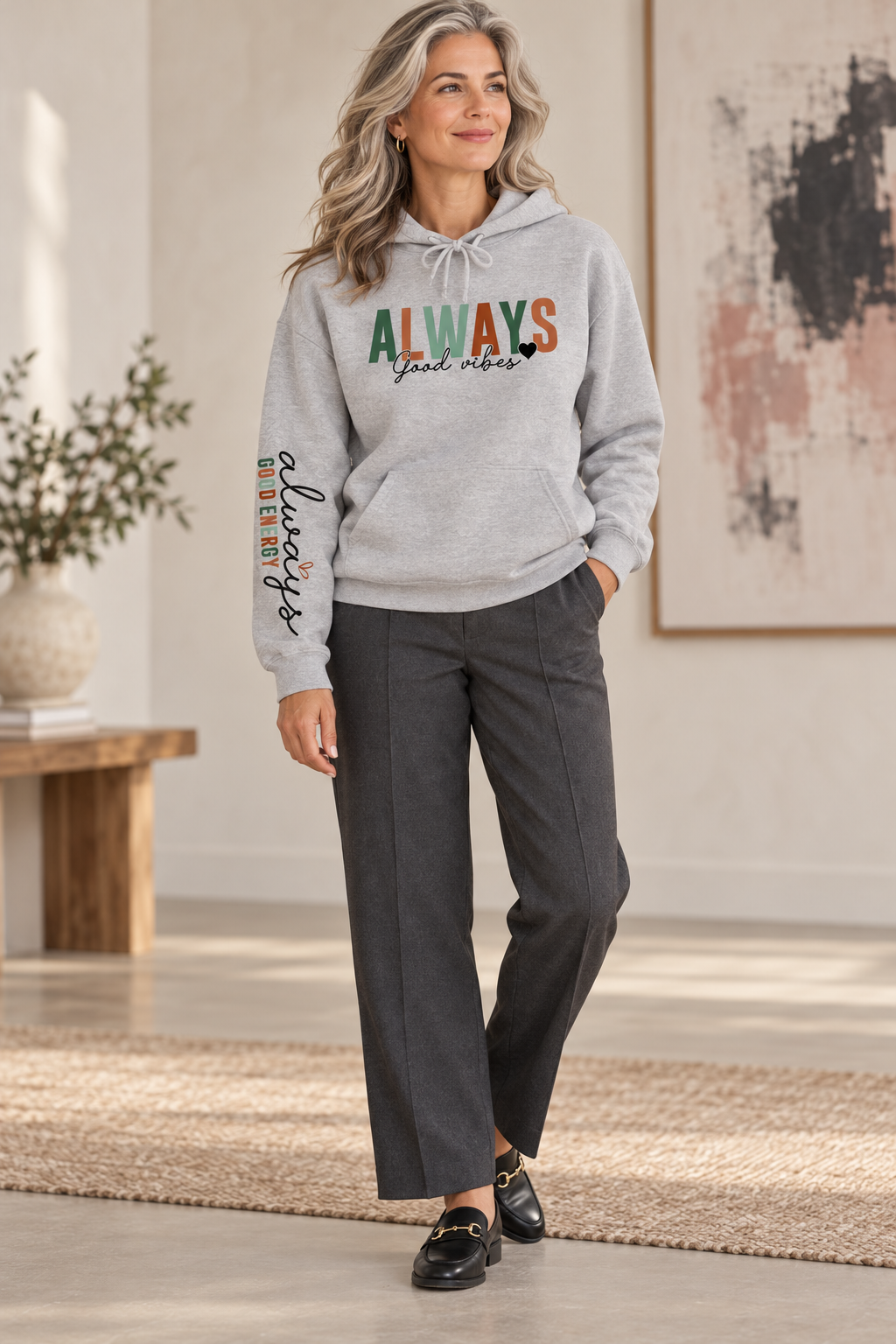 A woman stands in a bright indoor room wearing a light heather gray pullover hoodie with a front kangaroo pocket and drawstring hood, featuring the word 'ALWAYS' in block letters with 'Good vibes' in black script underneath with a solid small heart. The left sleeve has a vertical script graphic 'ALWAYS GOOD ENERGY', paired with dark charcoal wide-leg pants and black loafers.