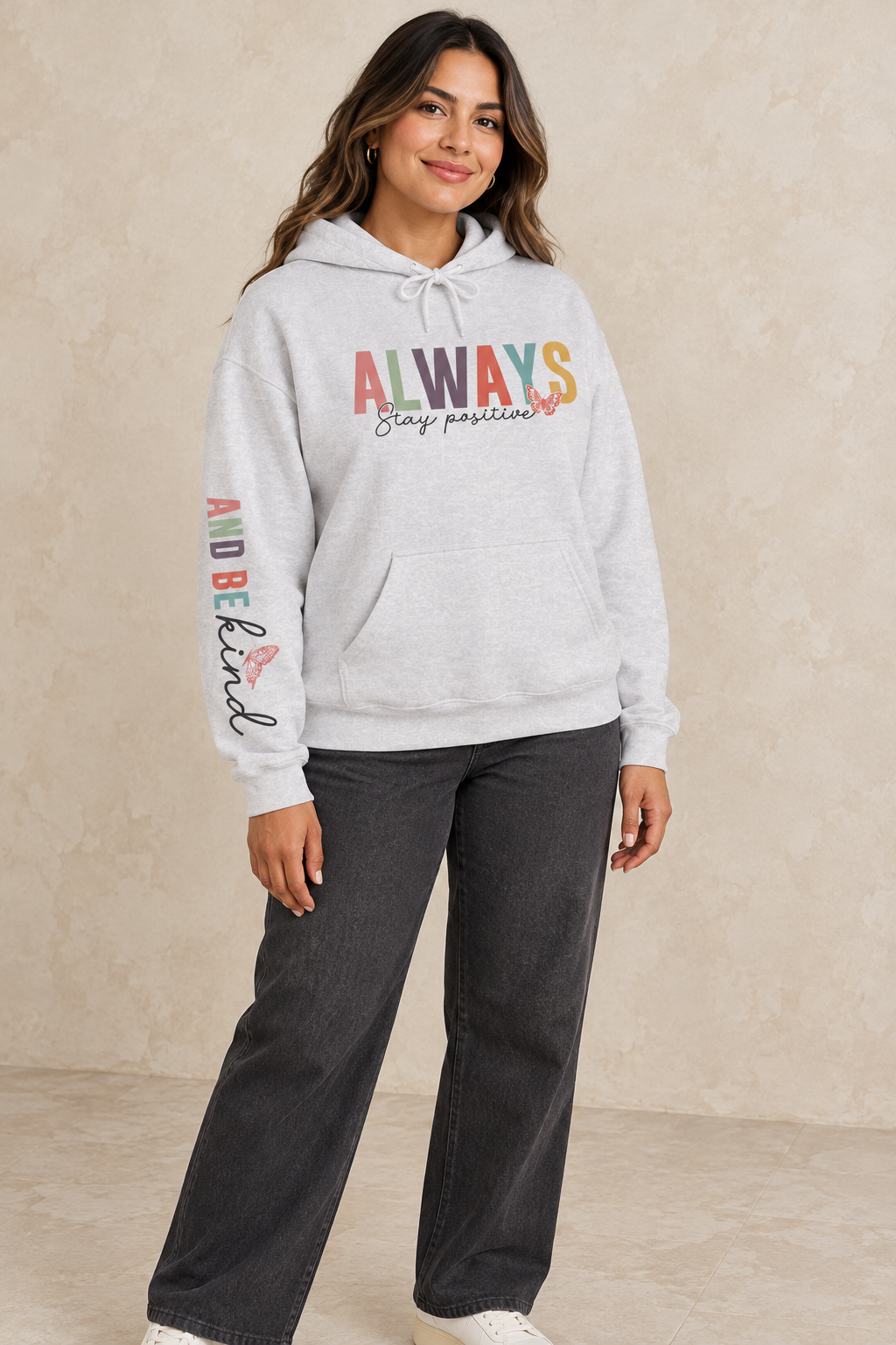 A woman wearing a light gray heather pullover hoodie featuring a colorful graphic on the chest that reads 'ALWAYS Stay positive' with small butterfly accents, and additional small graphics 'ALWAYS BE KIND' printed down the right sleeve, paired with dark gray wide-leg jeans against a textured beige studio backdrop.