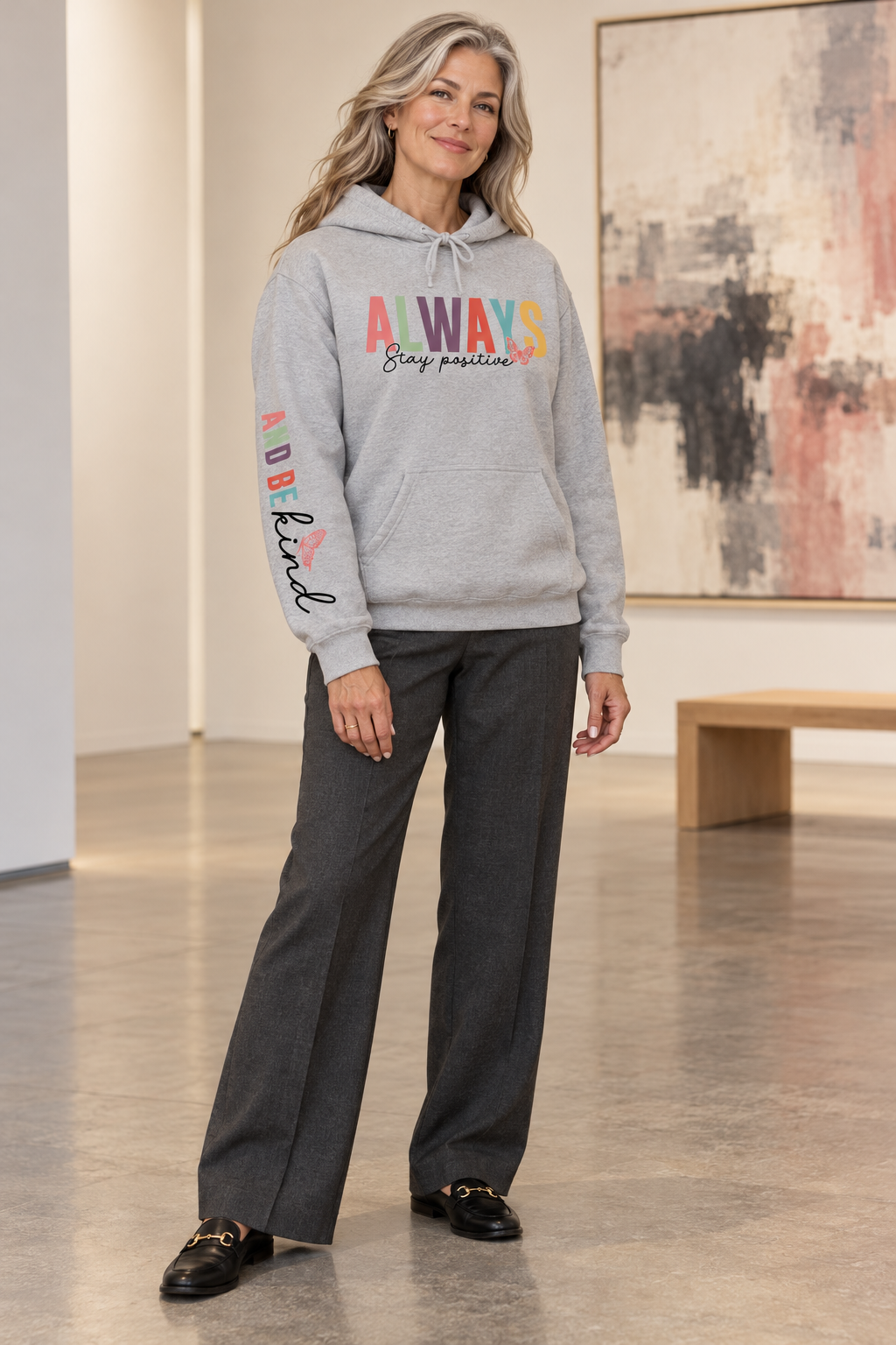A woman stands in a gallery wearing a light gray pullover hoodie featuring a colorful graphic on the chest that reads 'ALWAYS Stay positive' with small butterfly accents, and additional small graphics 'ALWAYS BE KIND' printed down the right sleeve, paired with dark gray wide-leg pants and black loafers.