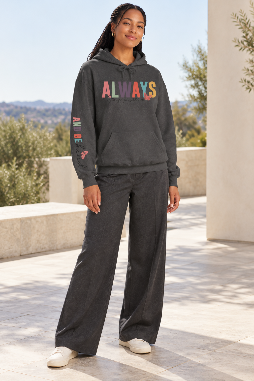 A woman modeling a dark gray pullover hoodie featuring a colorful graphic on the chest that reads 'ALWAYS Stay positive' with small butterfly accents, and additional small graphics 'ALWAYS BE KIND' printed down the right sleeve and matching wide-leg pants, standing outdoors on a patio.