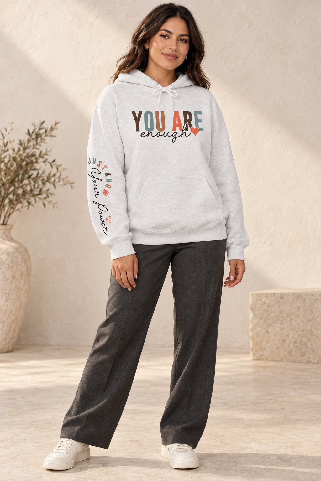 A woman modeling a light gray heathered hoodie with text graphics on the right sleeve 'Just Know Your Power', paired with blue jeans, shown from a side profile against a white background, paired with dark gray pants and white sneakers in a warm, sunlit indoor setting.