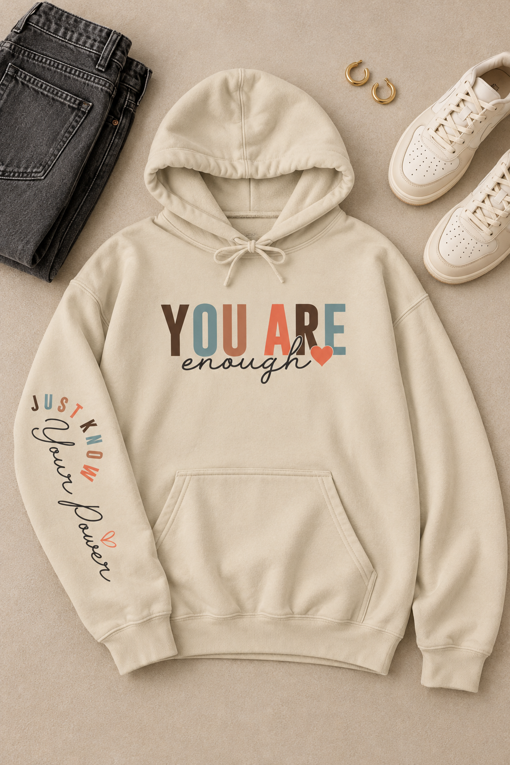 A beige hoodie with text graphics on the right sleeve 'Just Know Your Power', paired with blue jeans, shown from a side profile against a white background, styled in a flat lay with jeans, sneakers, and rings on a neutral background.