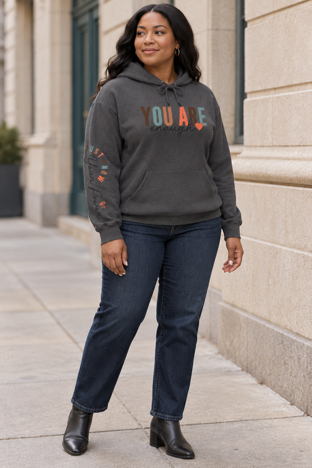 A woman wearing a dark gray hoodie with text graphics on the right sleeve 'Just Know Your Power', paired with blue jeans, shown from a side profile against a white background., paired with dark blue jeans and black ankle boots, standing on a city sidewalk.
