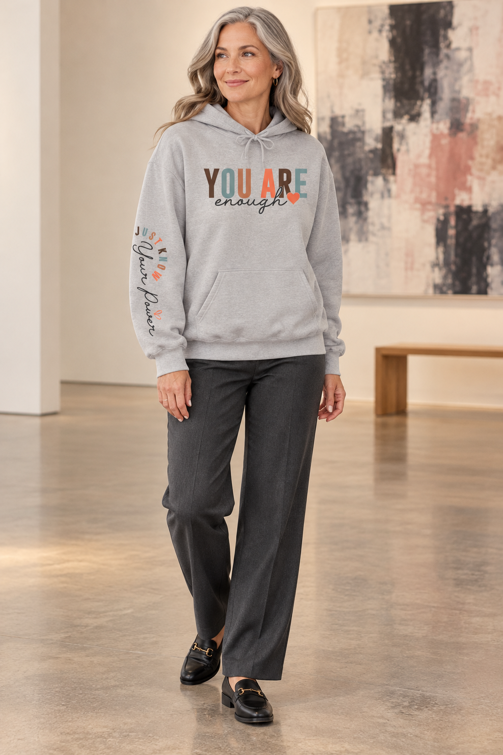 A woman modeling a light gray hoodie with text graphics on the right sleeve 'Just Know Your Power', paired with blue jeans, shown from a side profile against a white background, paired with dark gray pants and black loafers, standing in a gallery setting.