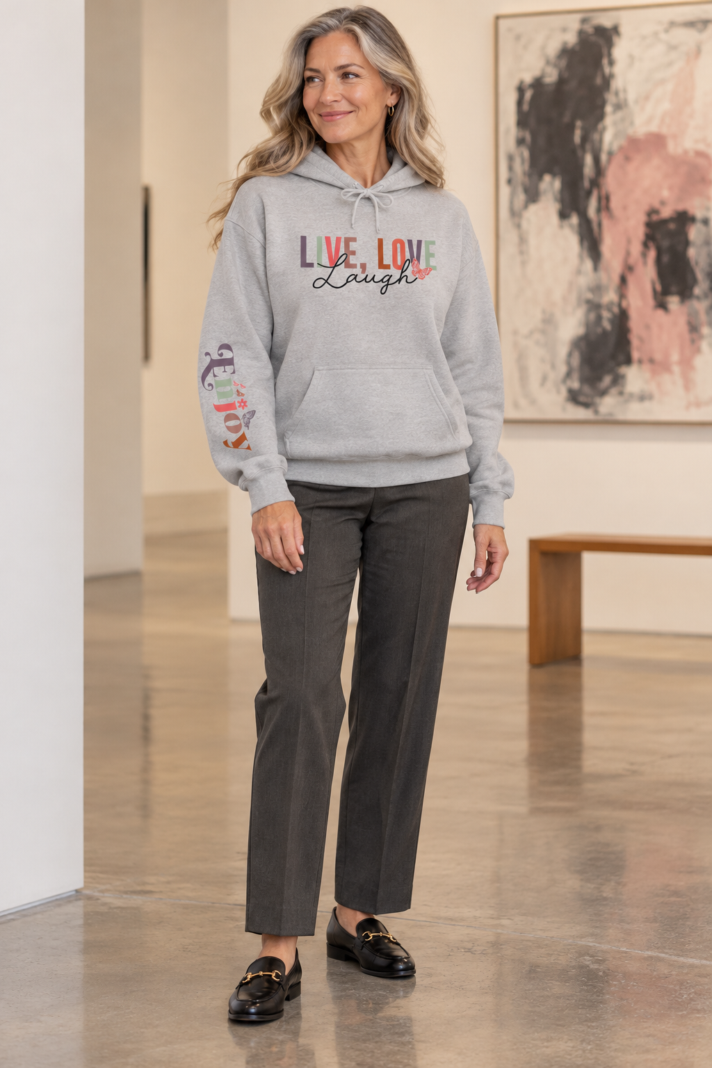 A woman modeling a light gray pullover hoodie with a colorful graphic on the chest reading 'LIVE, LOVE Laugh' and a small floral accent, plus a multicolor graphic on the right sleeve that reads 'ENJOY', paired with dark gray pants and black loafers in an indoor gallery setting.