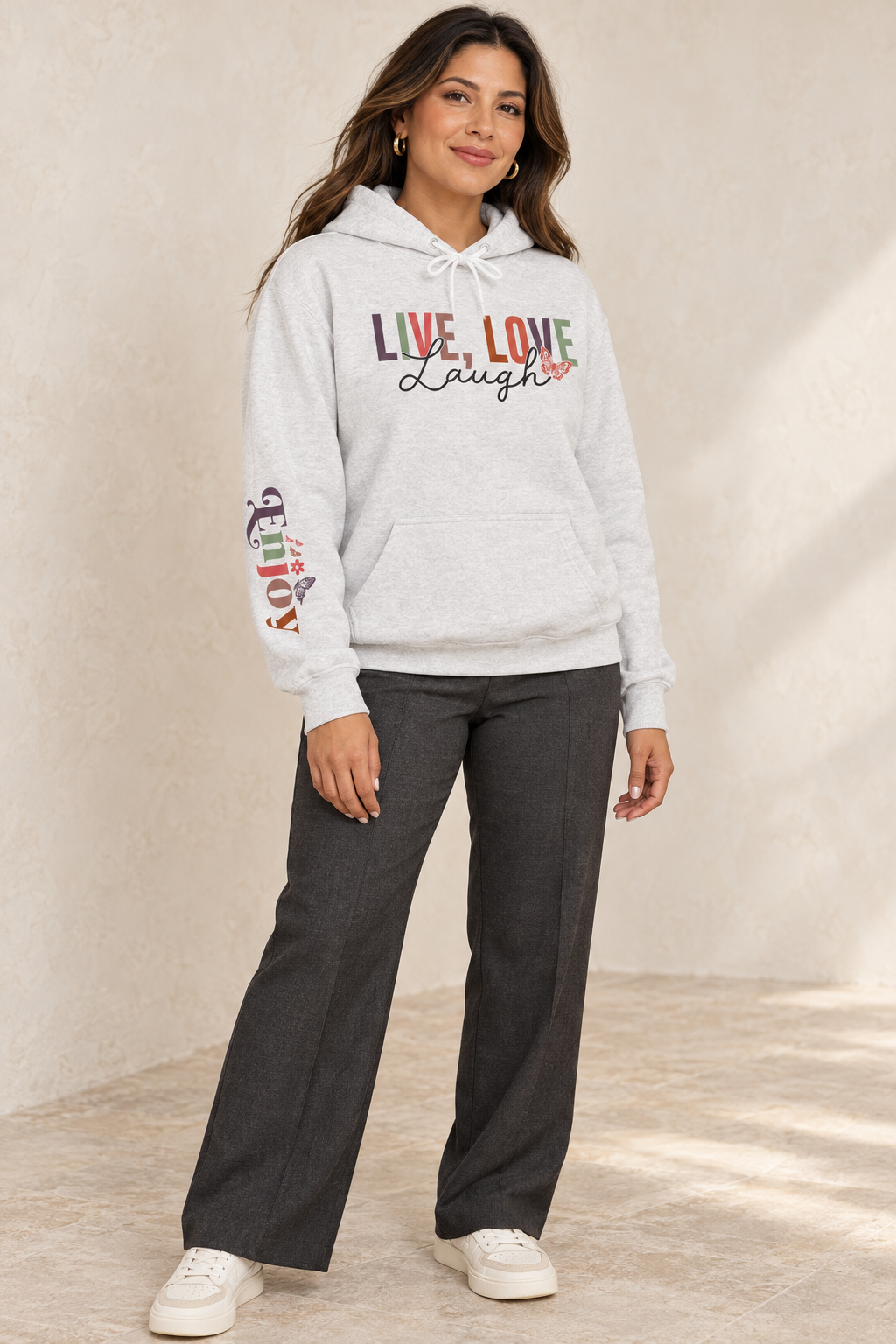 A woman modeling a light gray heather pullover hoodie with a colorful graphic on the chest reading 'LIVE, LOVE Laugh' and a small floral accent, plus a multicolor graphic on the right sleeve that reads 'ENJOY', paired with dark gray wide-leg pants and white sneakers, set against a textured beige wall.