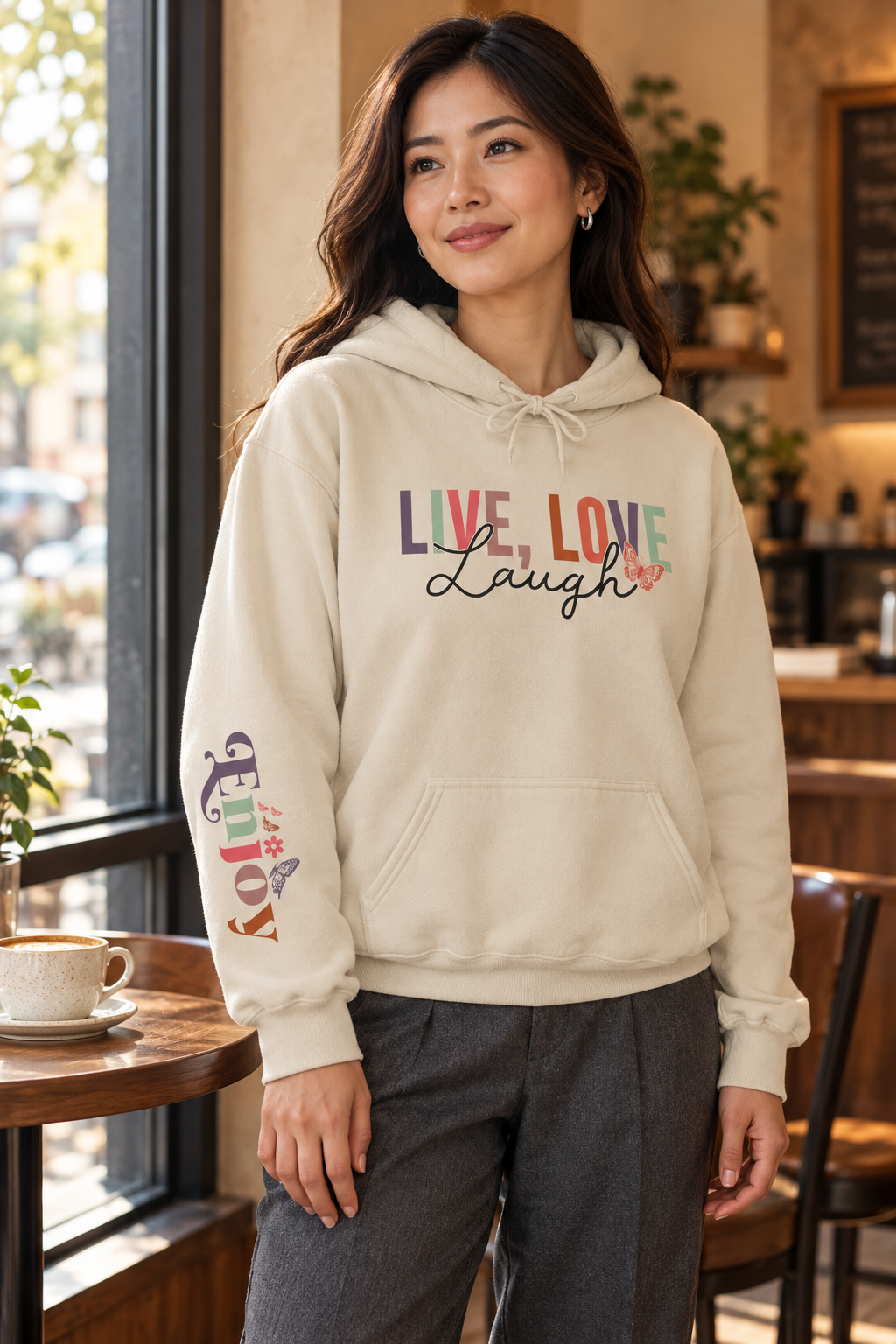A woman wearing a cream-colored pullover hoodie with a colorful graphic on the chest reading 'LIVE, LOVE Laugh' and a small floral accent, plus a multicolor graphic on the right sleeve that reads 'ENJOY', standing in a cozy cafe setting.