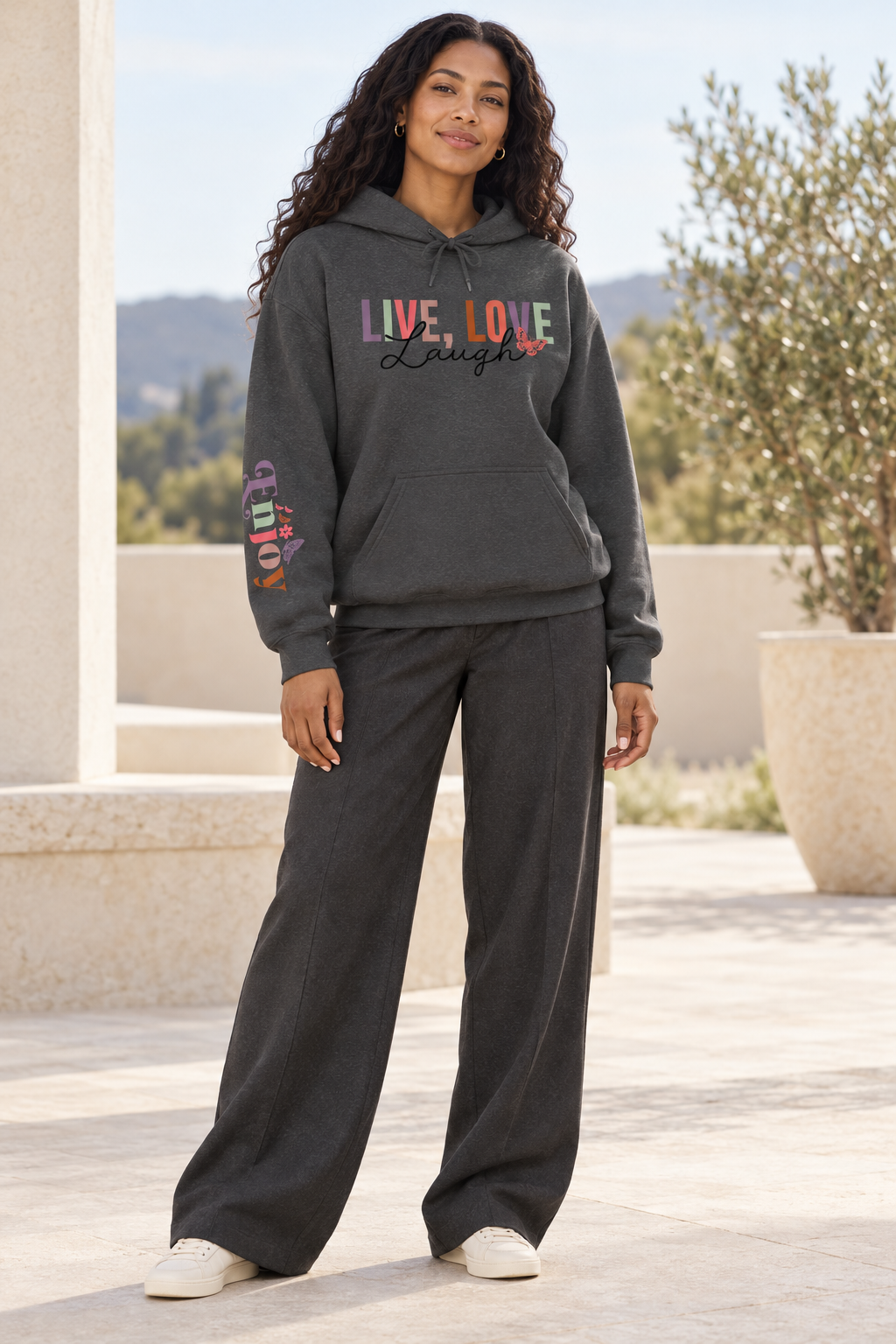 A woman modeling a dark gray pullover hoodie with a colorful graphic on the chest reading 'LIVE, LOVE Laugh' and a small floral accent, plus a multicolor graphic on the right sleeve that reads 'ENJOY' and matching wide-leg sweatpants, standing outdoors on a patio.