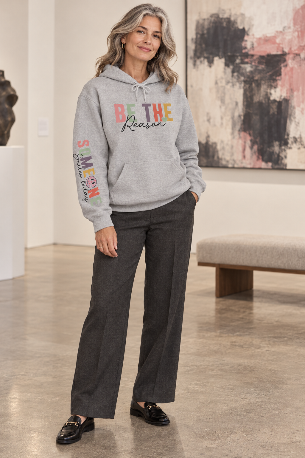 A woman modeling a light gray pullover hoodie with a colorful graphic on the chest reading 'BE THE Reason' and 'Someone Smiles today' with a smiley face printed down the right sleeve, paired with dark charcoal wide-leg pants and black loafers in an indoor gallery setting.