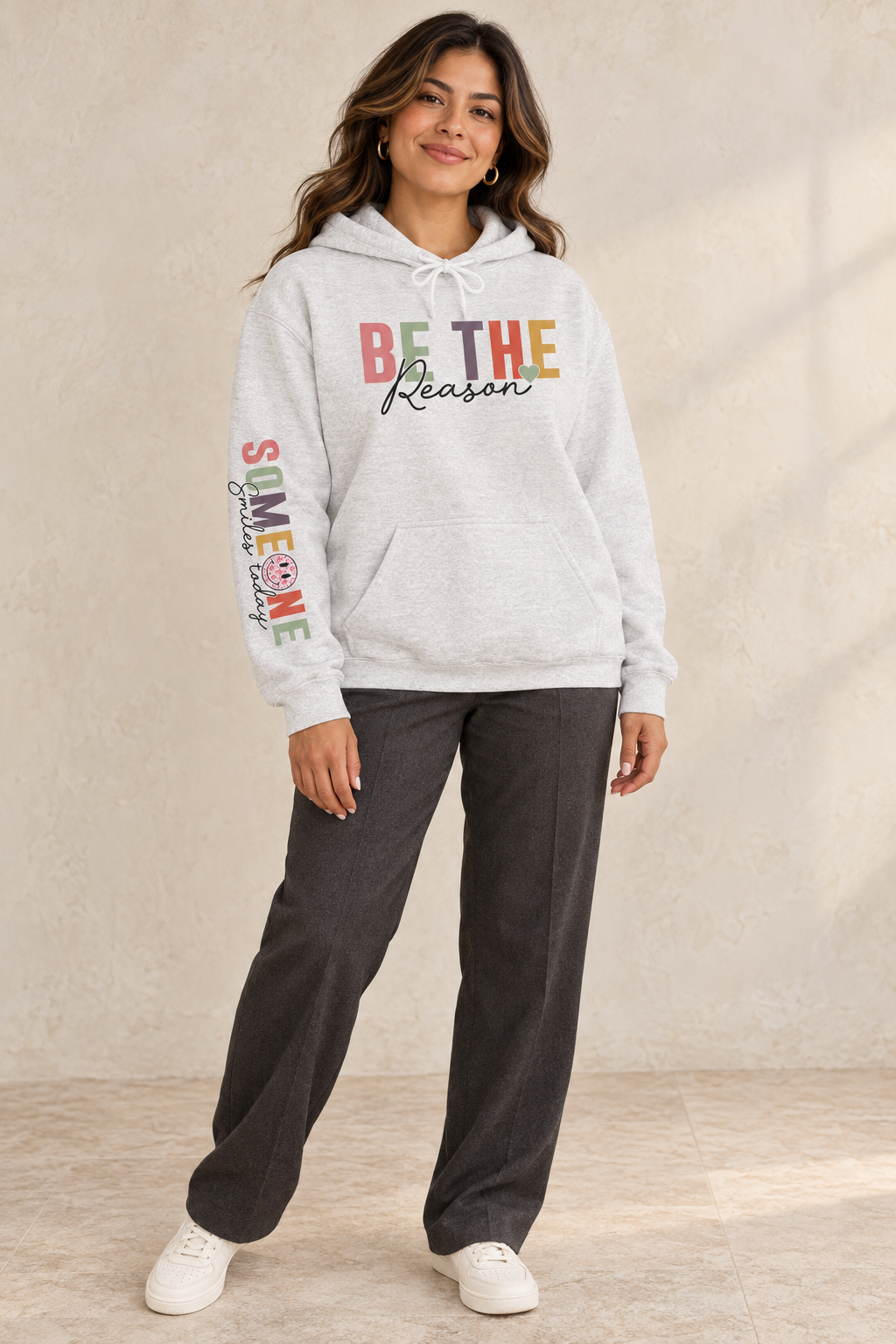 A woman modeling a light gray pullover hoodie with a colorful graphic on the chest reading 'BE THE Reason' and 'Someone Smiles today' with a smiley face printed down the right sleeve, paired with dark gray sweatpants and white sneakers against a textured beige wall.
