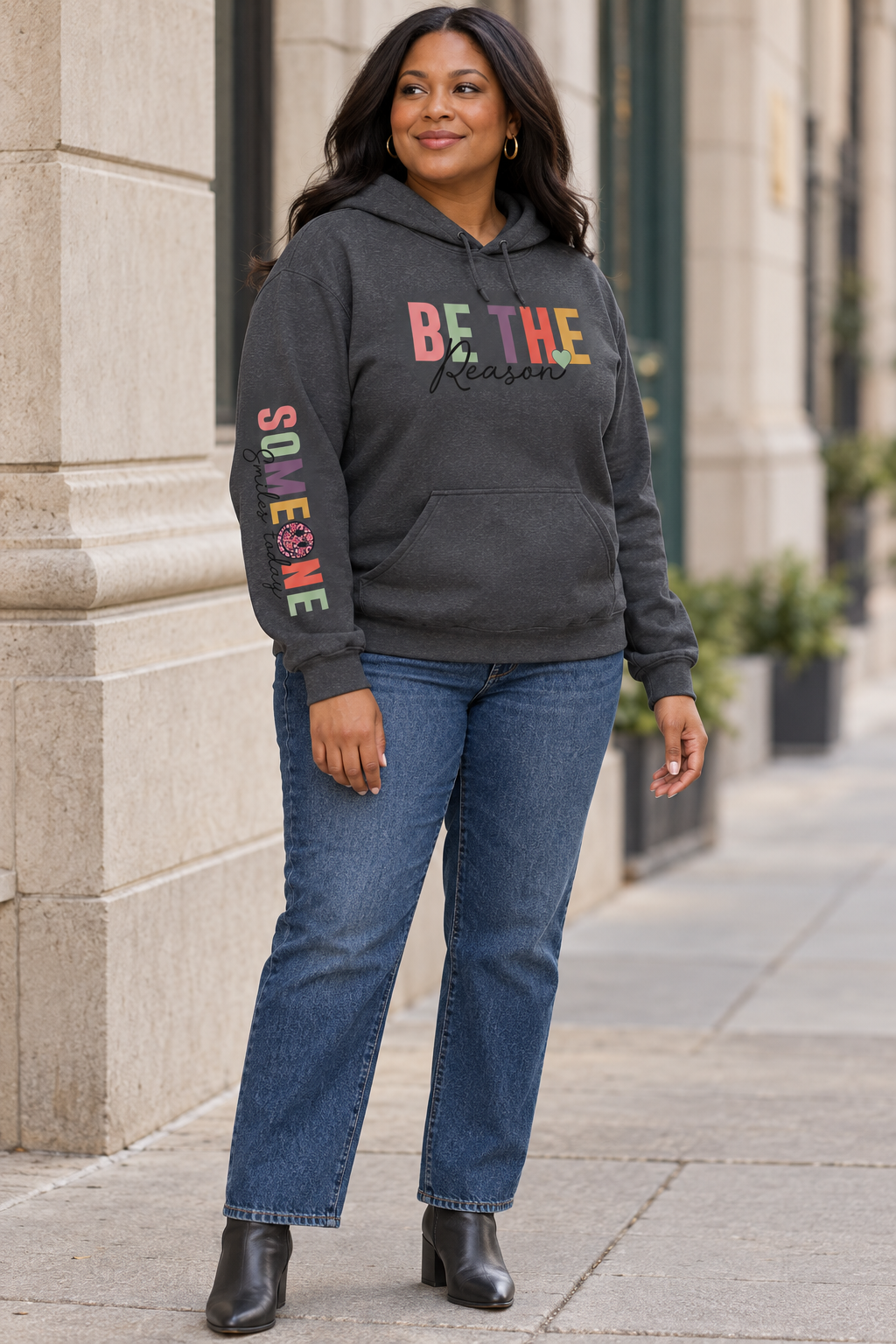 A woman wearing a dark gray pullover hoodie with a colorful graphic on the chest reading 'BE THE Reason' and 'Someone Smiles today' with a smiley face printed down the right sleeve, paired with blue jeans and black ankle boots, standing on a city sidewalk.