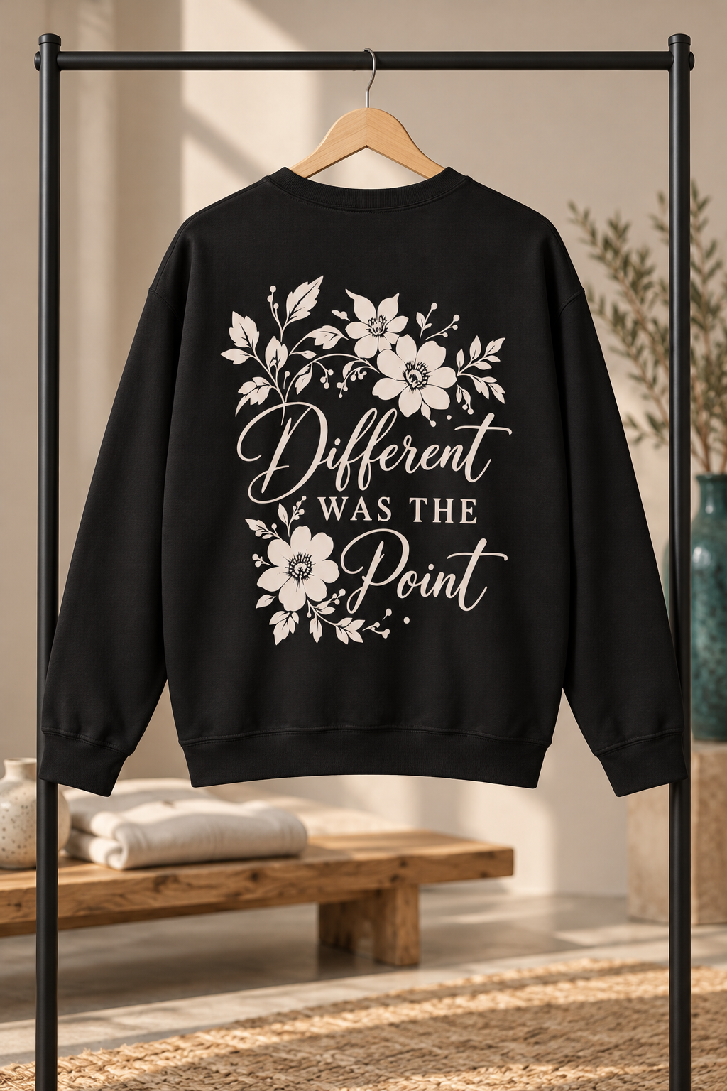 A black crewneck sweatshirt hanging on a wooden hanger from a black clothing rack, featuring a cream graphic print on the back with floral illustrations and the text 'Different WAS THE Point'. The background is a warm, softly lit room with a wooden bench and neutral decor.