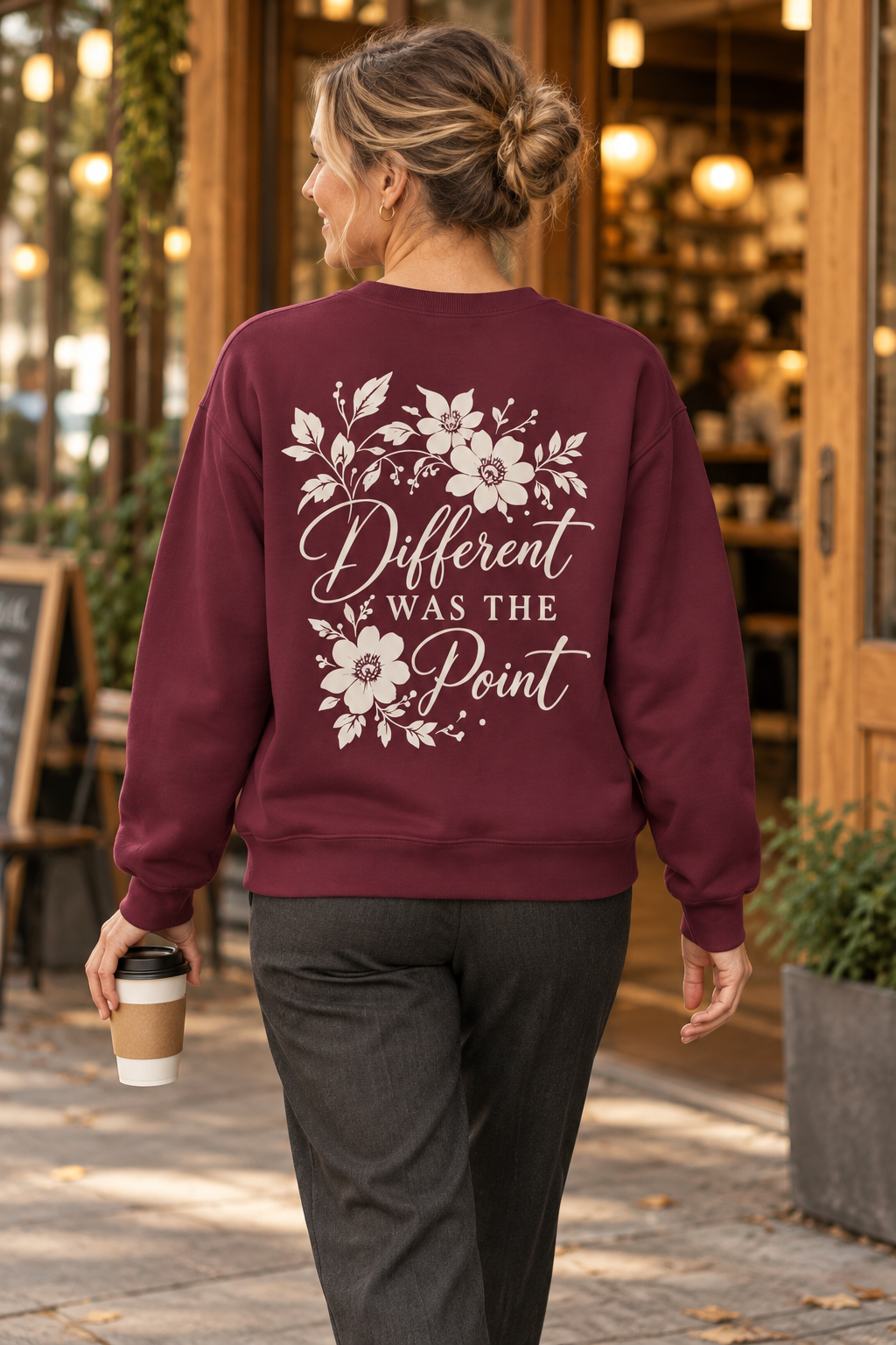 A woman viewed from behind wearing a maroon crewneck sweatshirt with a cream floral graphic and the text 'Different WAS THE Point' printed on the back. She is walking outdoors in front of a warm, softly blurred storefront.