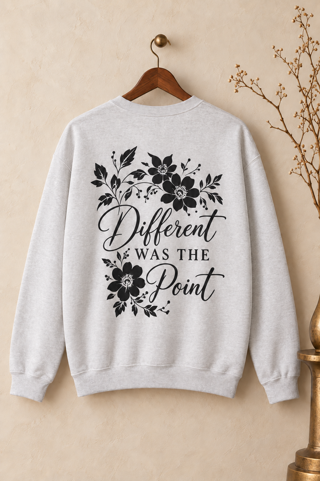 A light heather gray crewneck sweatshirt hanging on a wooden hanger against a warm beige wall. The back of the sweatshirt features a black graphic print with floral illustrations and the text 'Different WAS THE Point' in a mix of script and serif fonts.