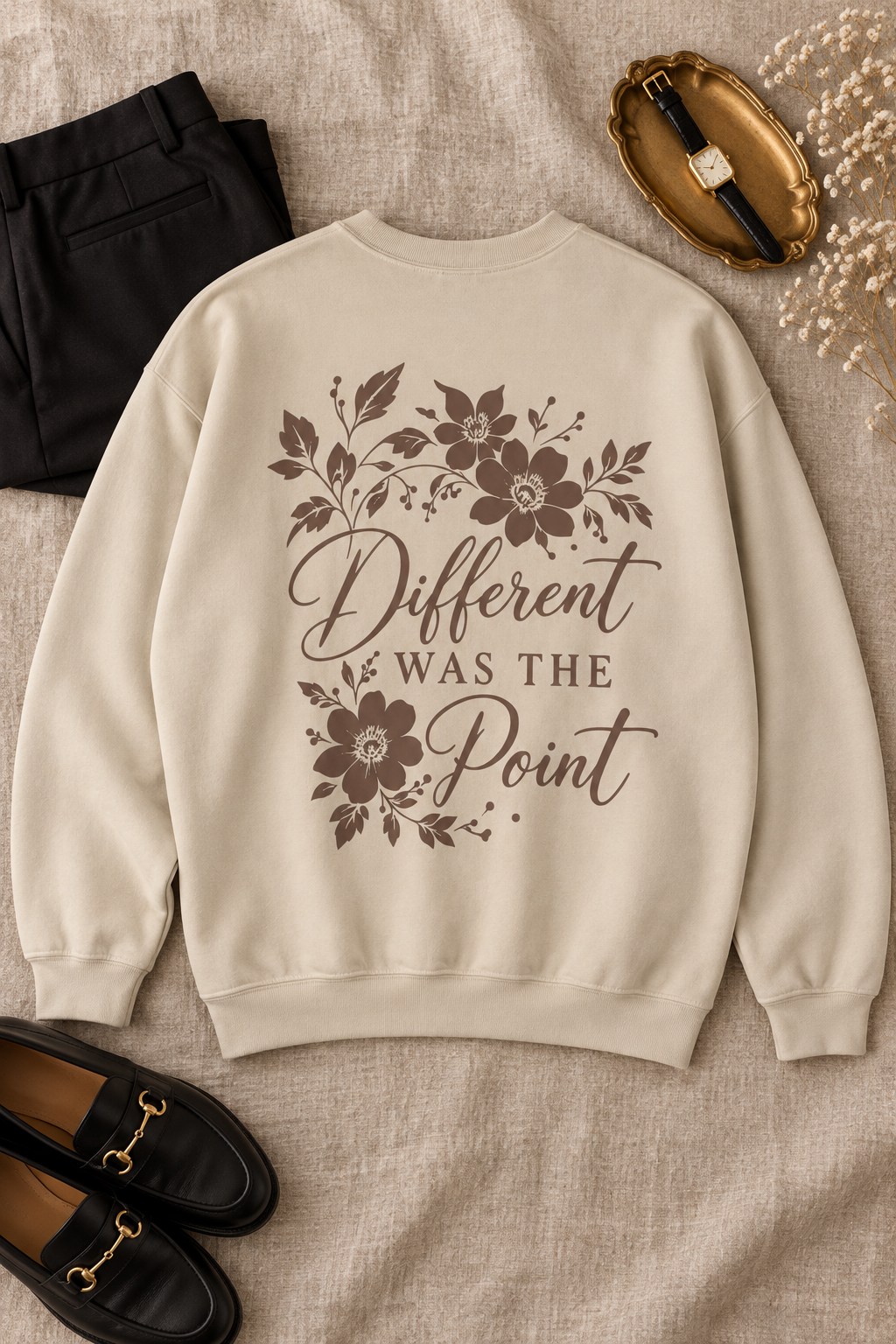 A beige crewneck sweatshirt with a brown floral graphic and the text 'Different WAS THE Point' printed on the back, styled in a flat lay with black pants, black loafers, a gold watch, and dried flowers on a beige linen background.