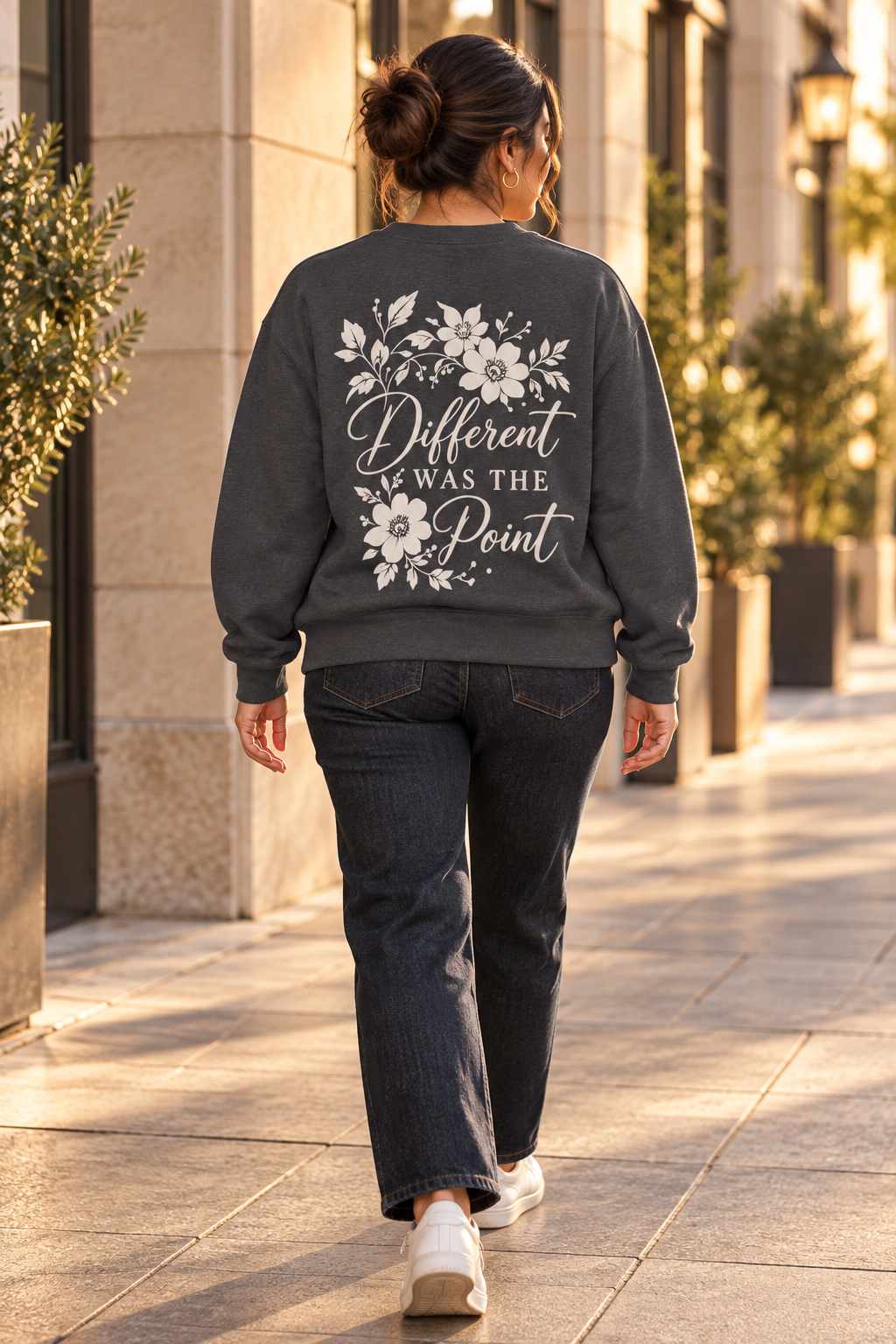 A woman viewed from behind walking on a sunlit city sidewalk, wearing a dark charcoal gray sweatshirt with a cream floral graphic and the phrase 'Different Was The Point' across the back, paired with dark jeans and white sneakers.