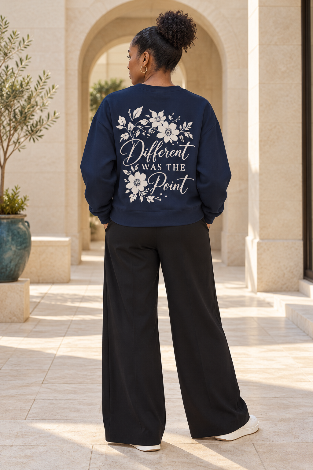 A woman stands outdoors in a sunny stone walkway, viewed from the back. She is wearing a navy blue sweatshirt with a large white floral graphic and the phrase 'Different WAS THE Point' printed on the back, paired with black wide-leg pants and white sneakers.